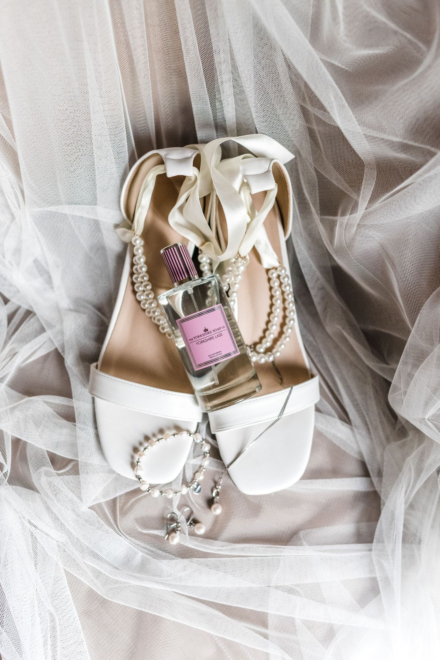 Wedding accessories including white sandals, perfume bottle, and pearl jewelry on tulle fabric.