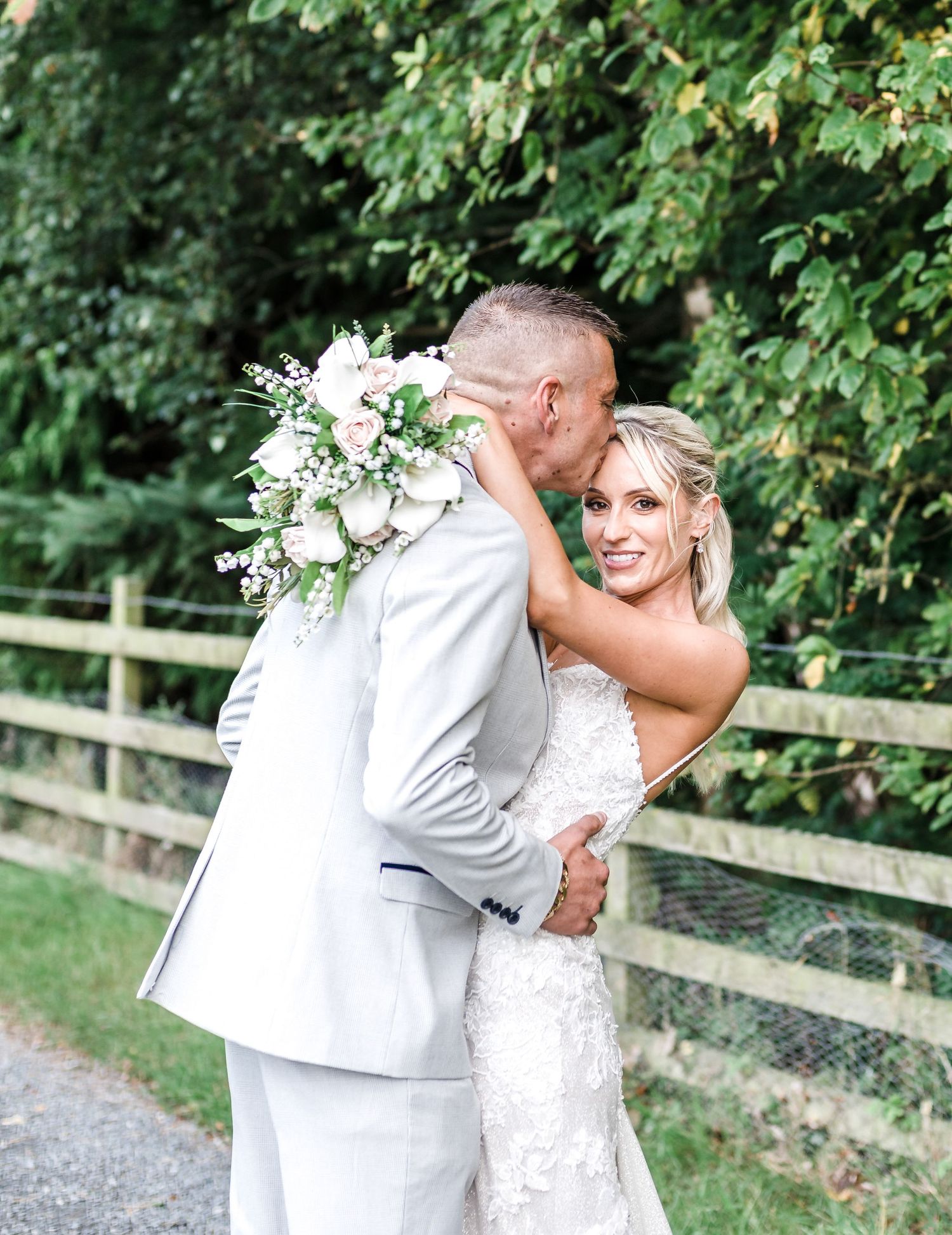 A romantic wedding couple embraces outdoors by a wooden fence surrounded by lush greenery.