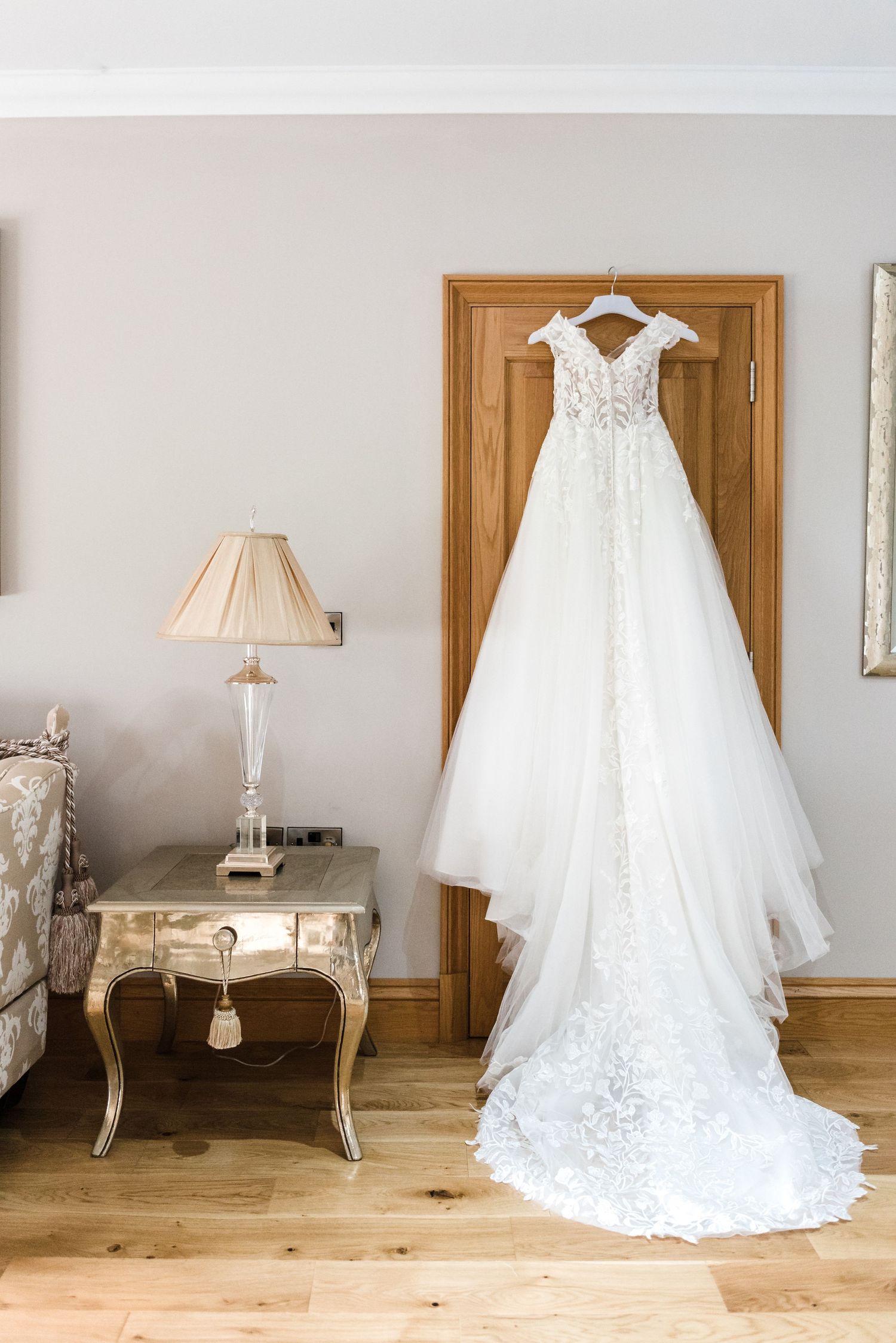 Elegant white lace wedding dress hanging on wooden door with vintage furniture nearby.
