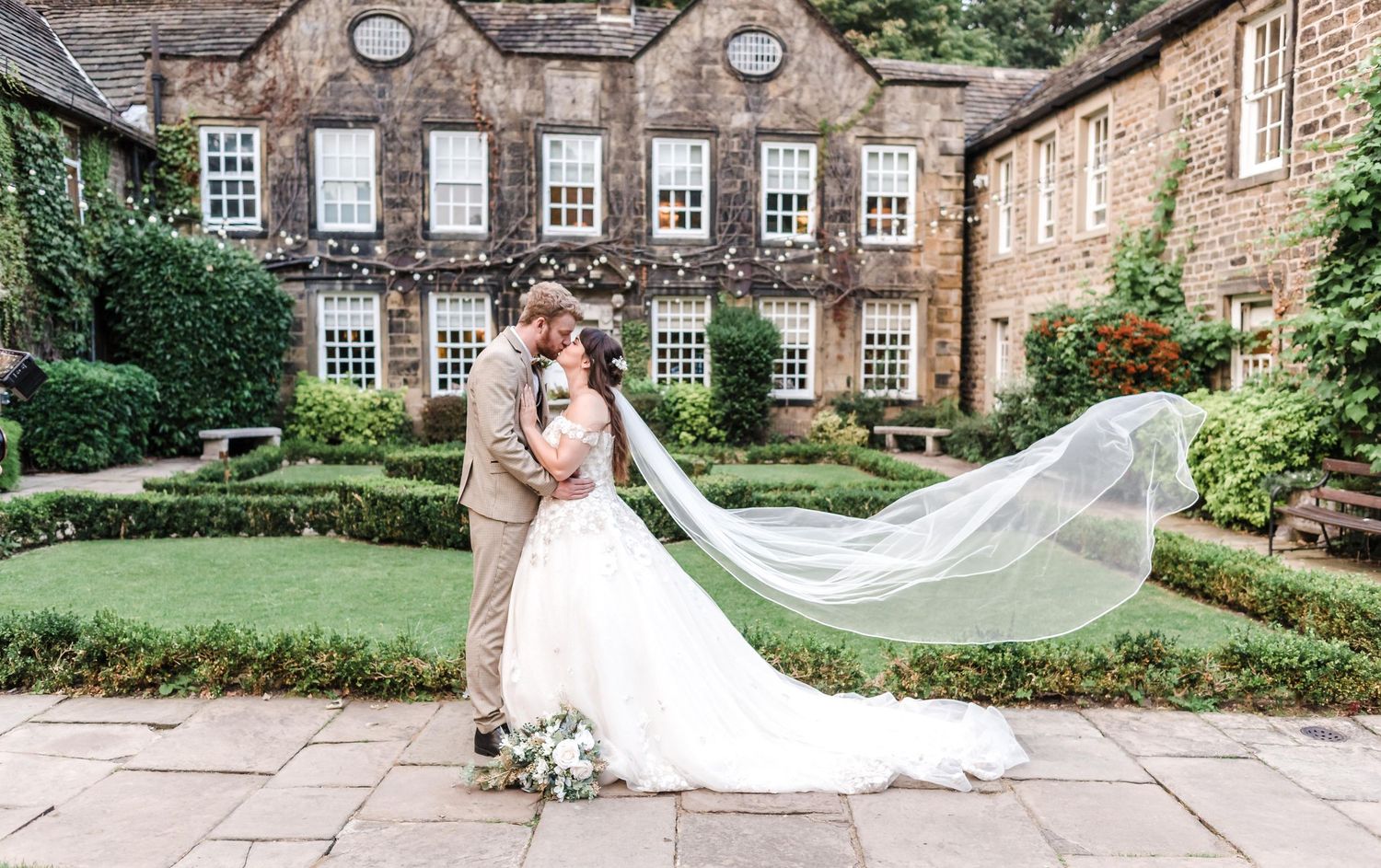 Wedding couple embracing outside historic mansion with flowing veil in garden setting.
