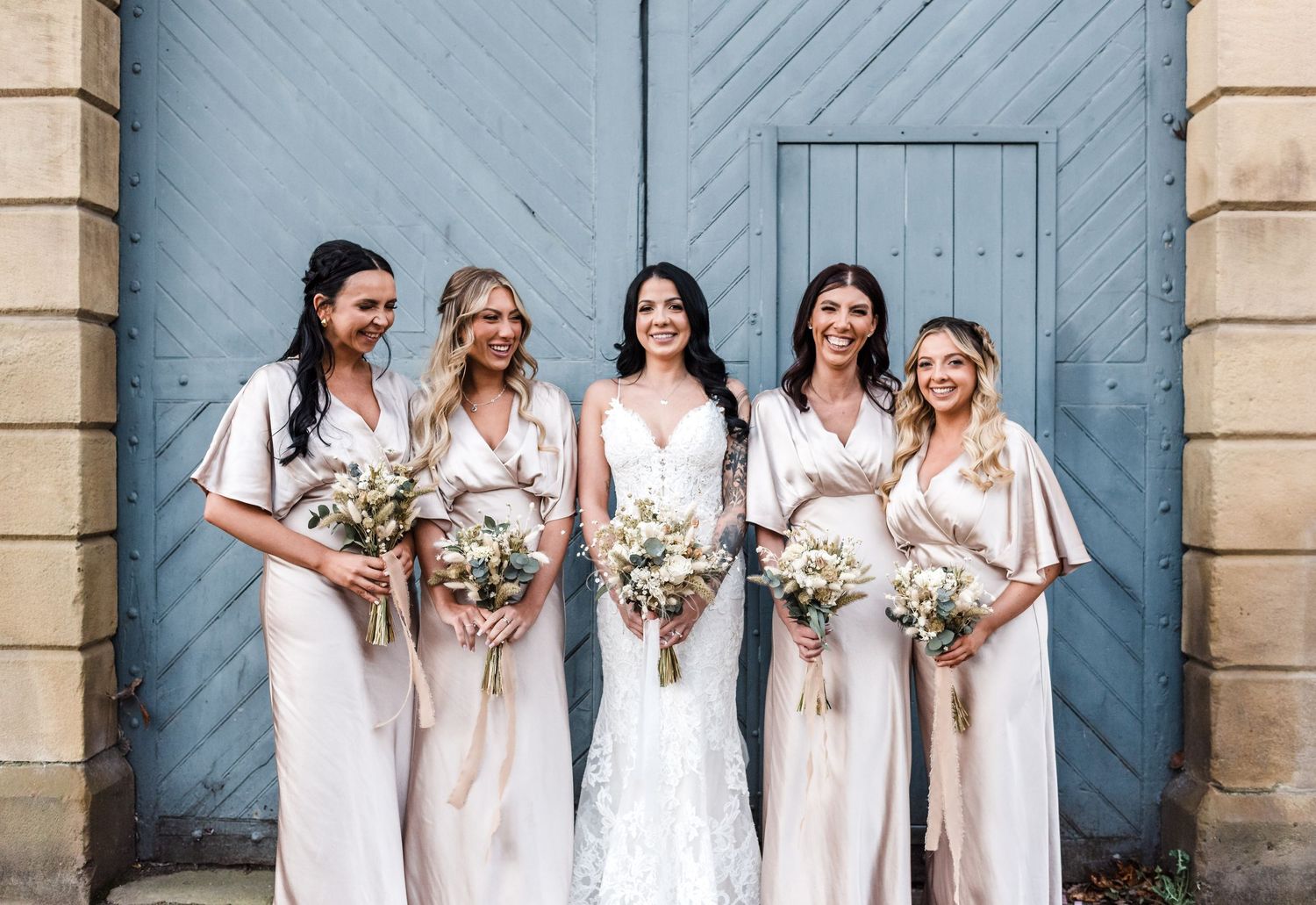 Bridal party in light colored dresses posing against blue wooden door.