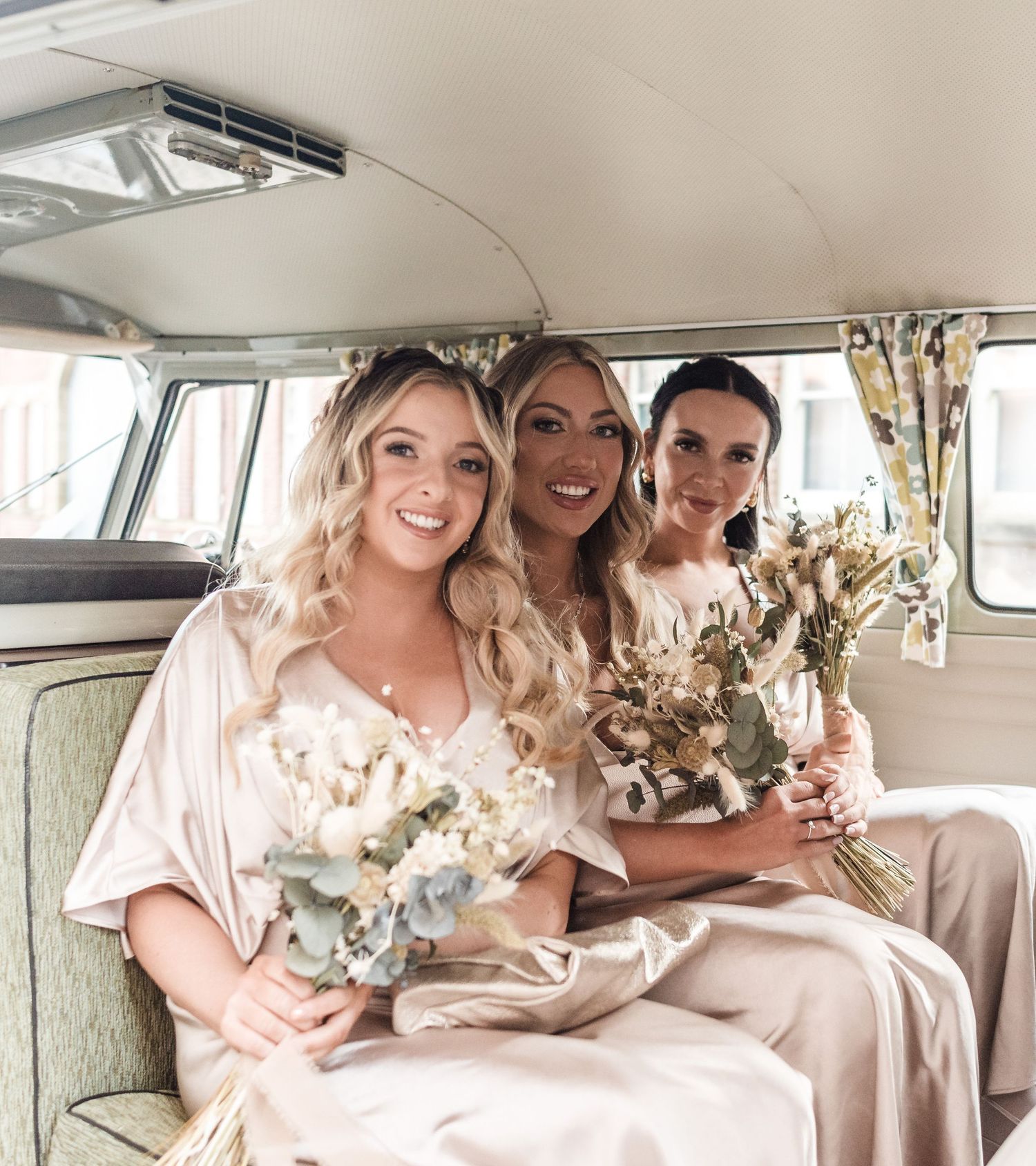 Three attendants in silk dresses pose with bouquets inside vintage van.