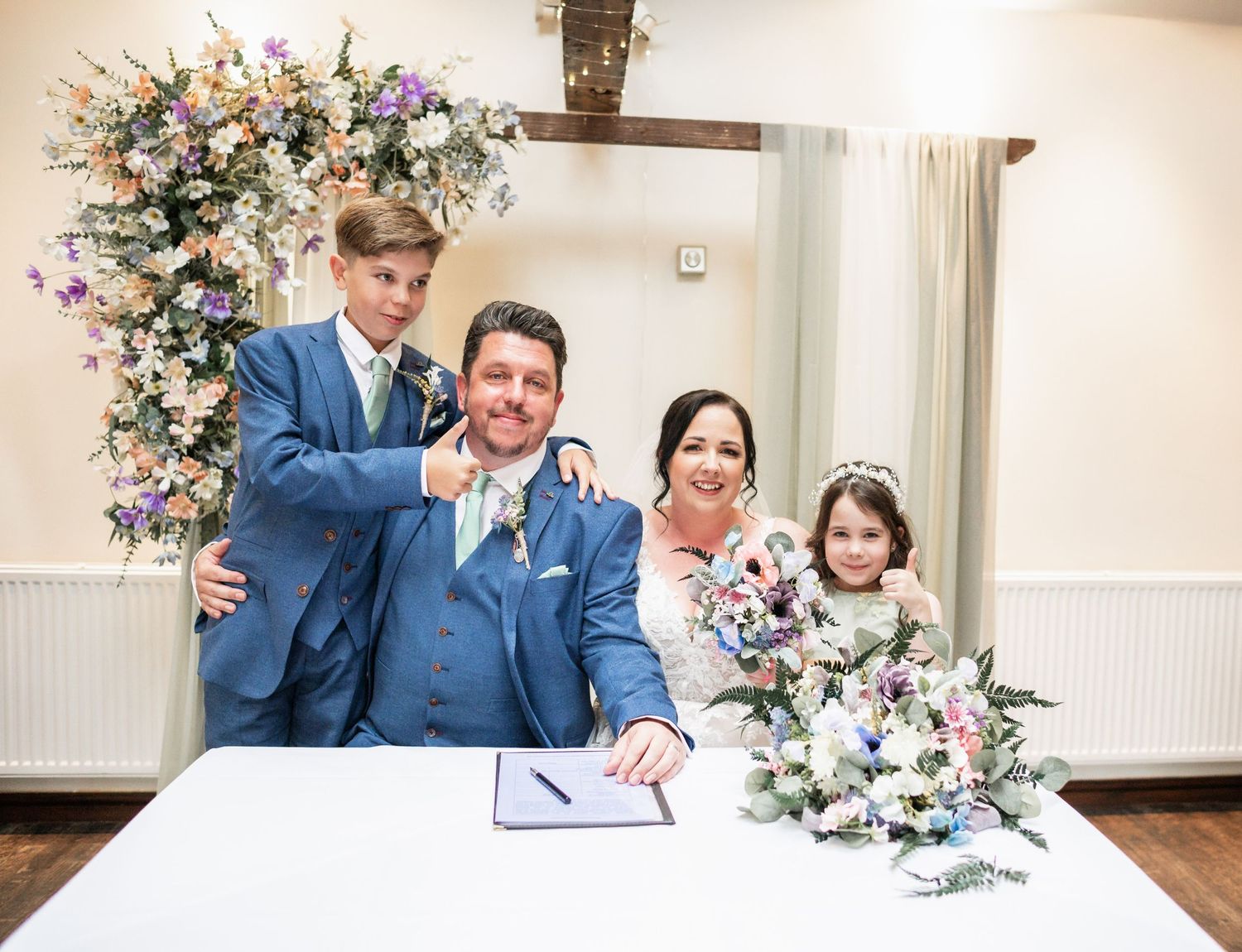 Wedding party gathered around signing table with floral arch and white decor in bright indoor venue.