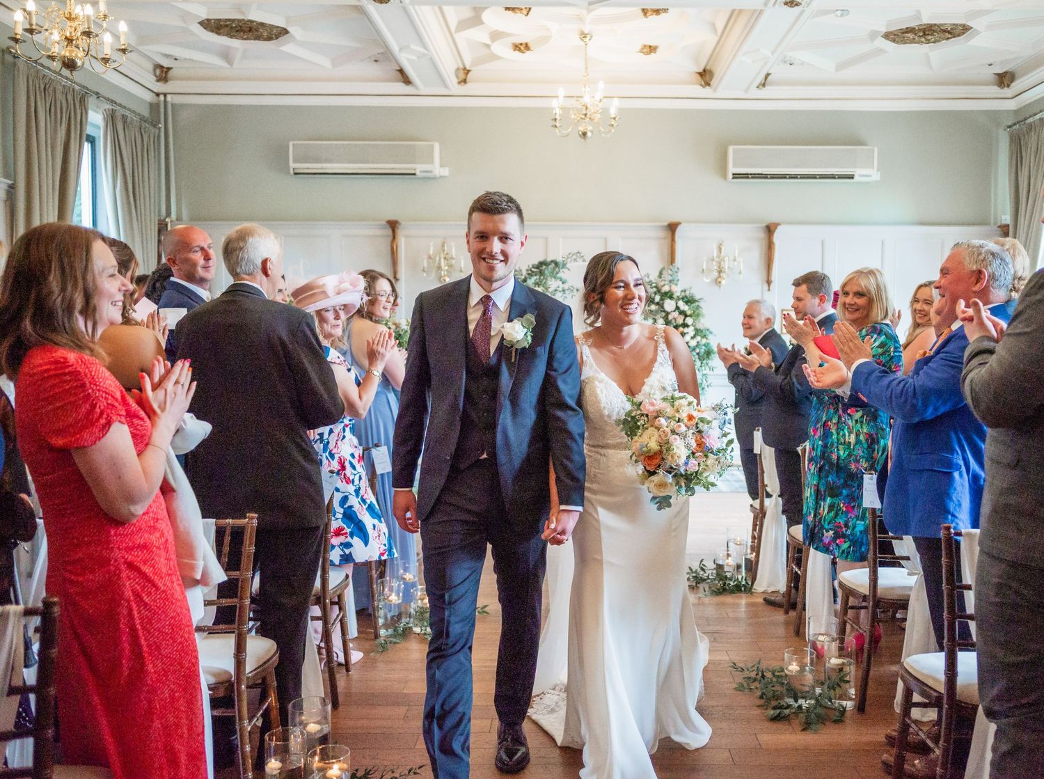 Wedding ceremony processional in an elegant room with chandeliers and decorative floral aisle markers.
