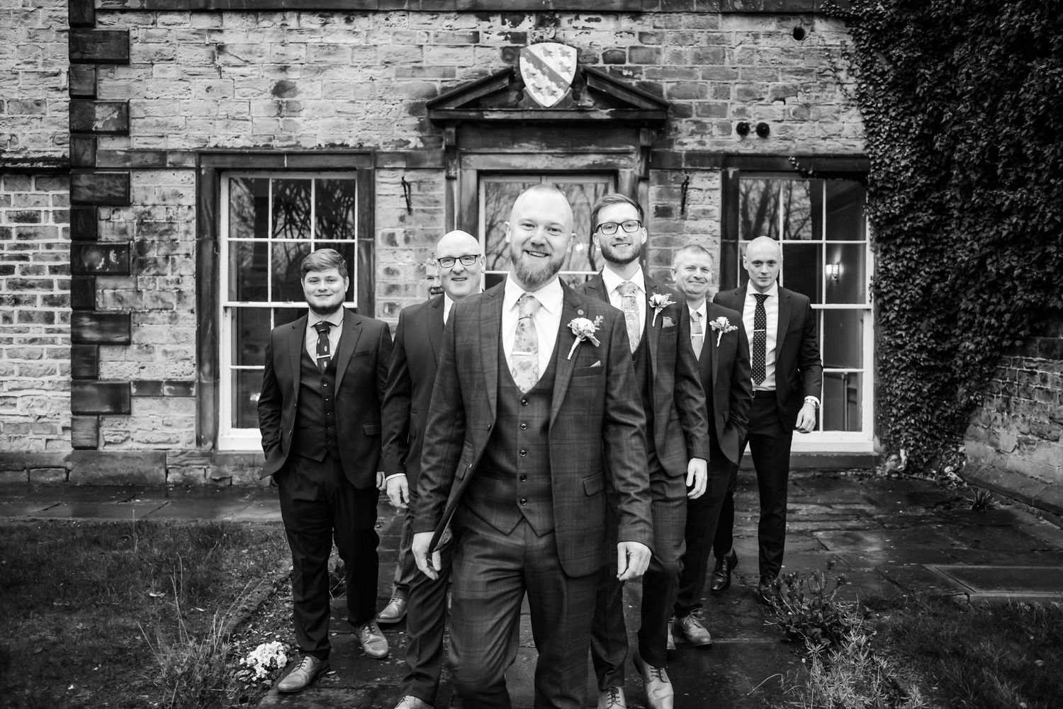 Black and white wedding party photo with groomsmen in formal attire standing in front of a stone building.