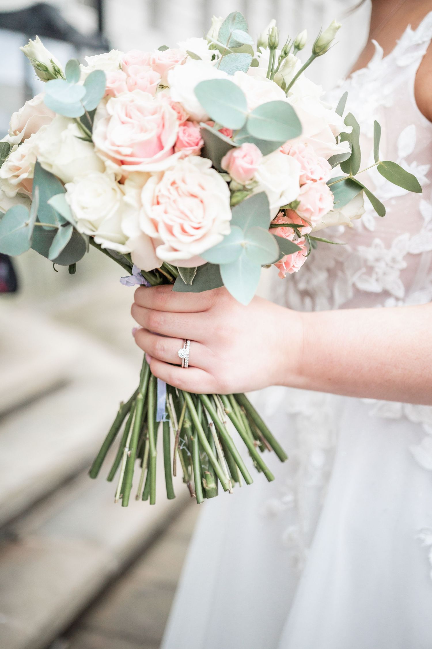 A bridal bouquet of pink roses and eucalyptus held by hands wearing a wedding ring.