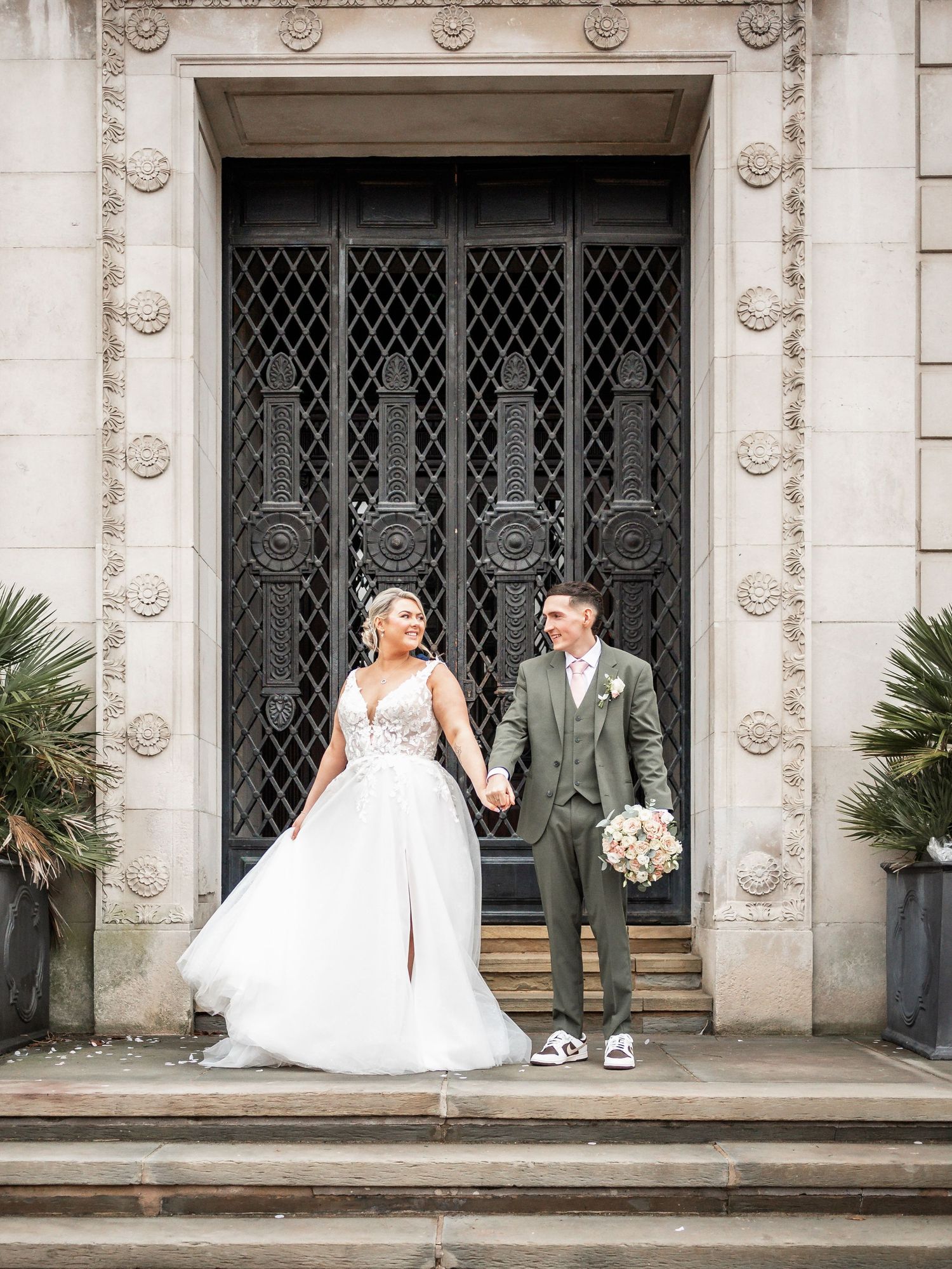 Newlyweds pose together on stone steps in front of decorative iron gate entrance.