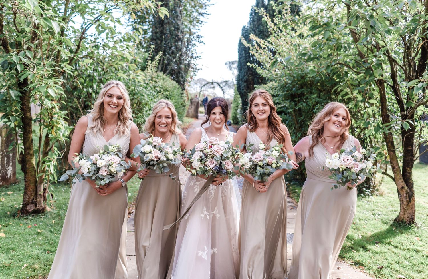 Wedding party in neutral dresses poses with floral bouquets in an outdoor garden setting.
