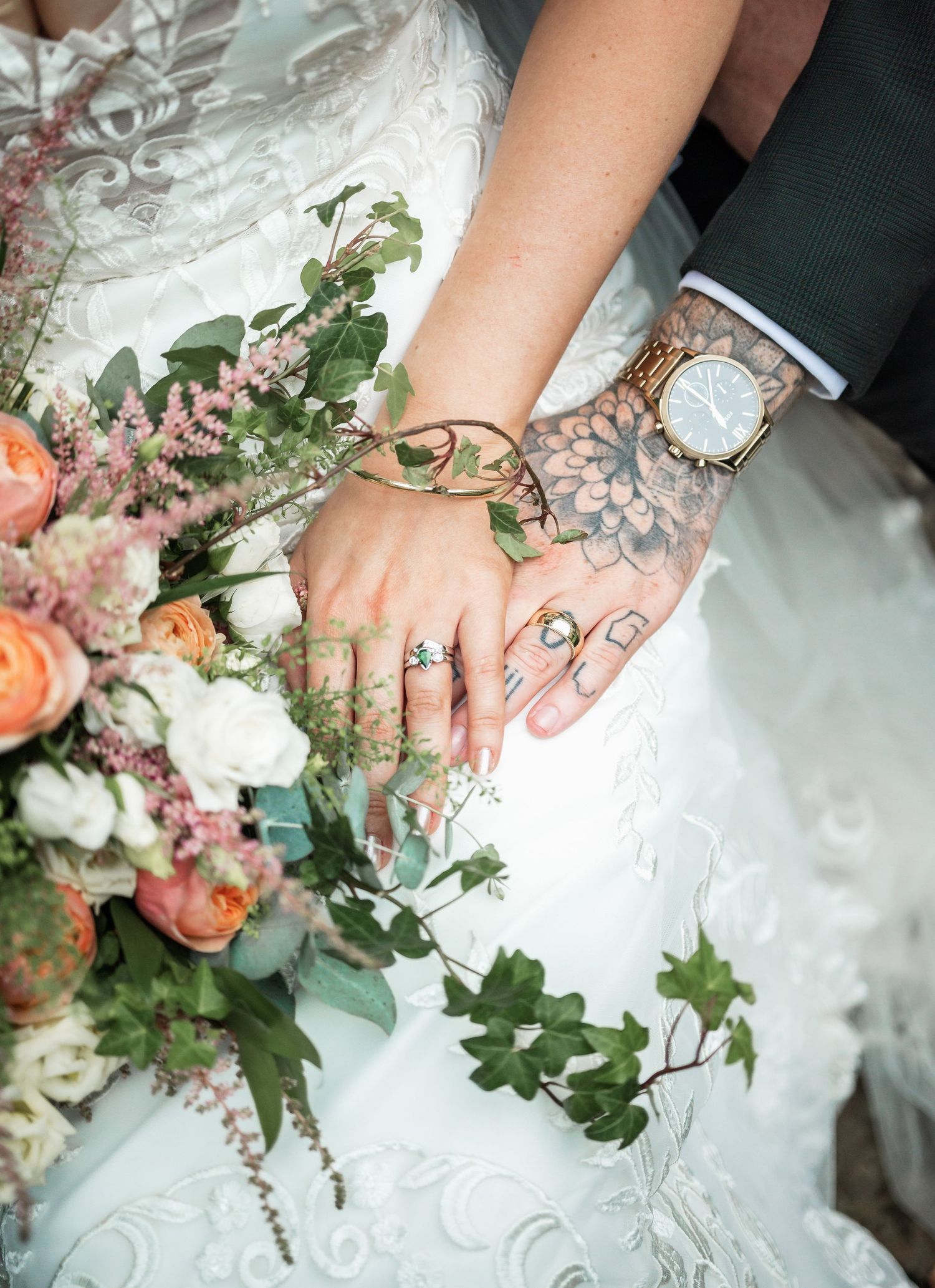 Close-up of wedding rings and floral details with coral roses and delicate greenery against white lace.