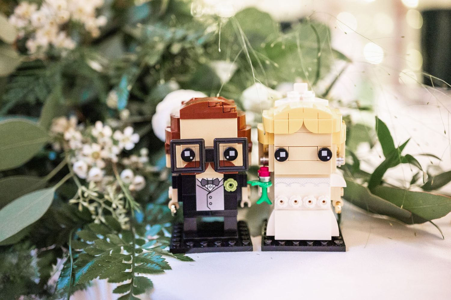 Cute LEGO bride and groom cake toppers displayed with greenery and white flowers.