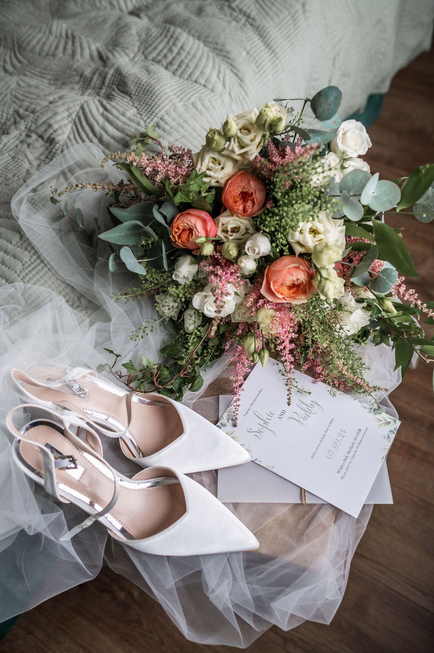 Bridal details with coral and white bouquet, white heels, and tulle fabric on wooden floor.