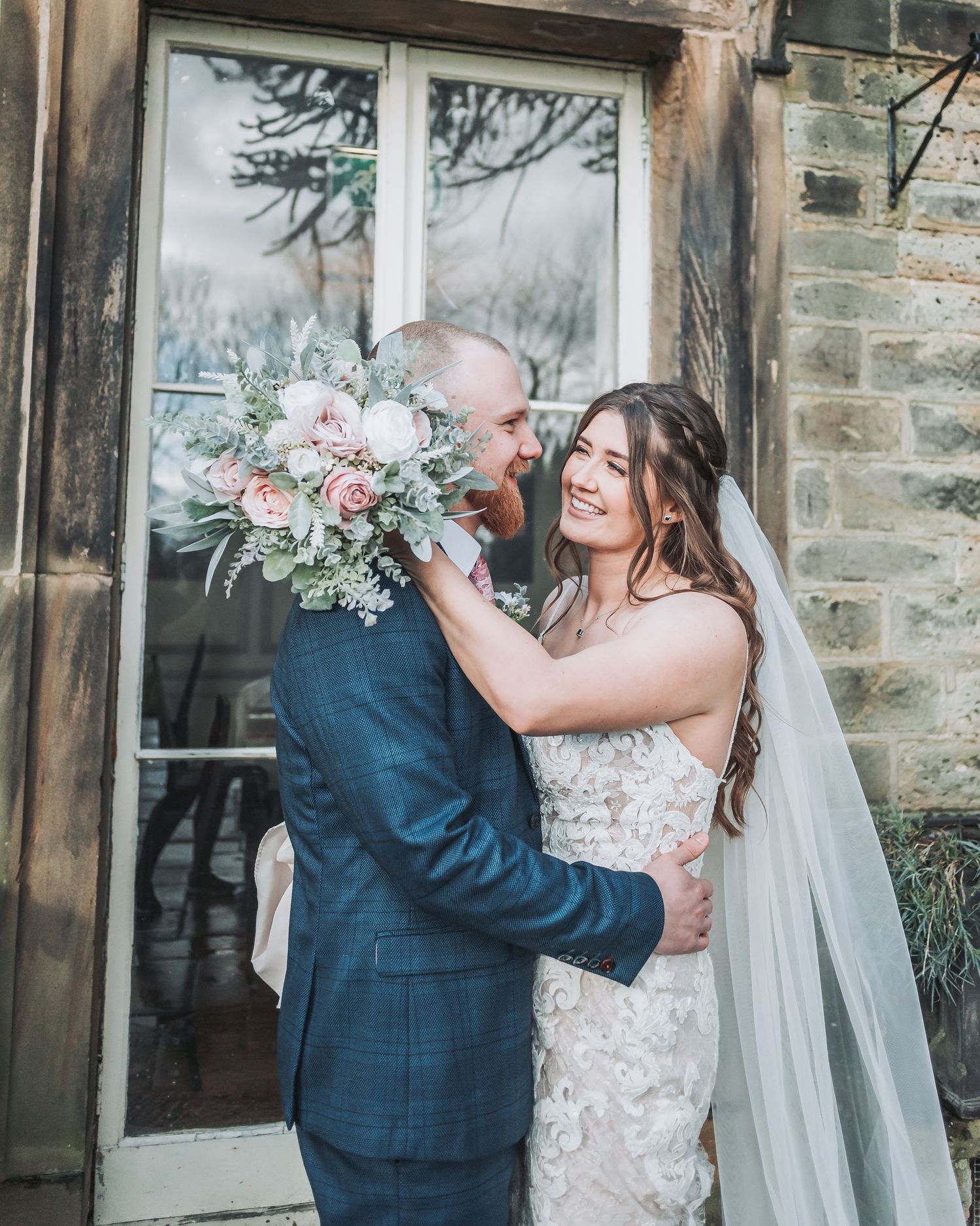 Joyful wedding moment captured with pink and white bouquet against stone building backdrop.