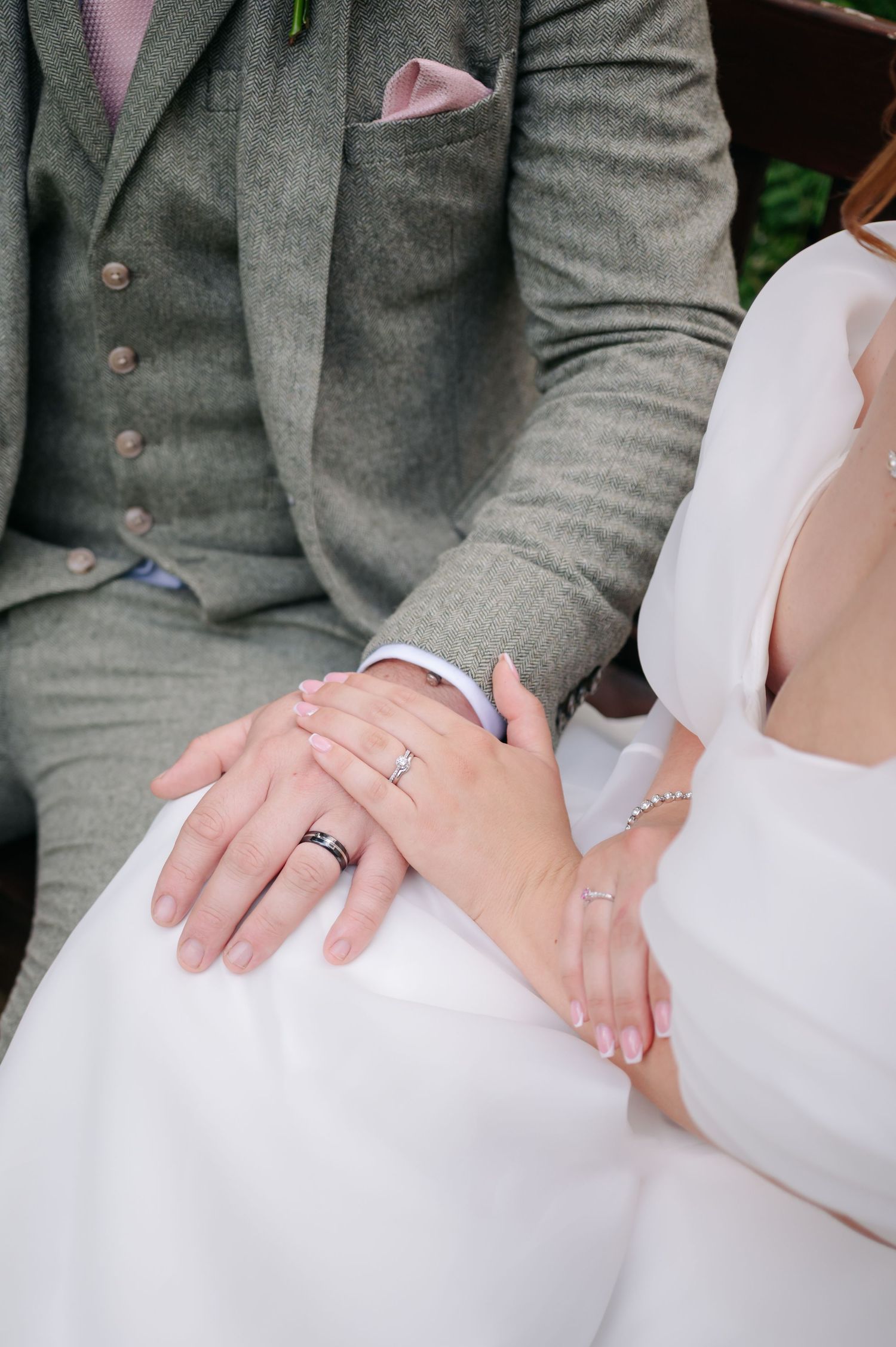 A close-up shot of hands with wedding rings against a gray suit and white dress.