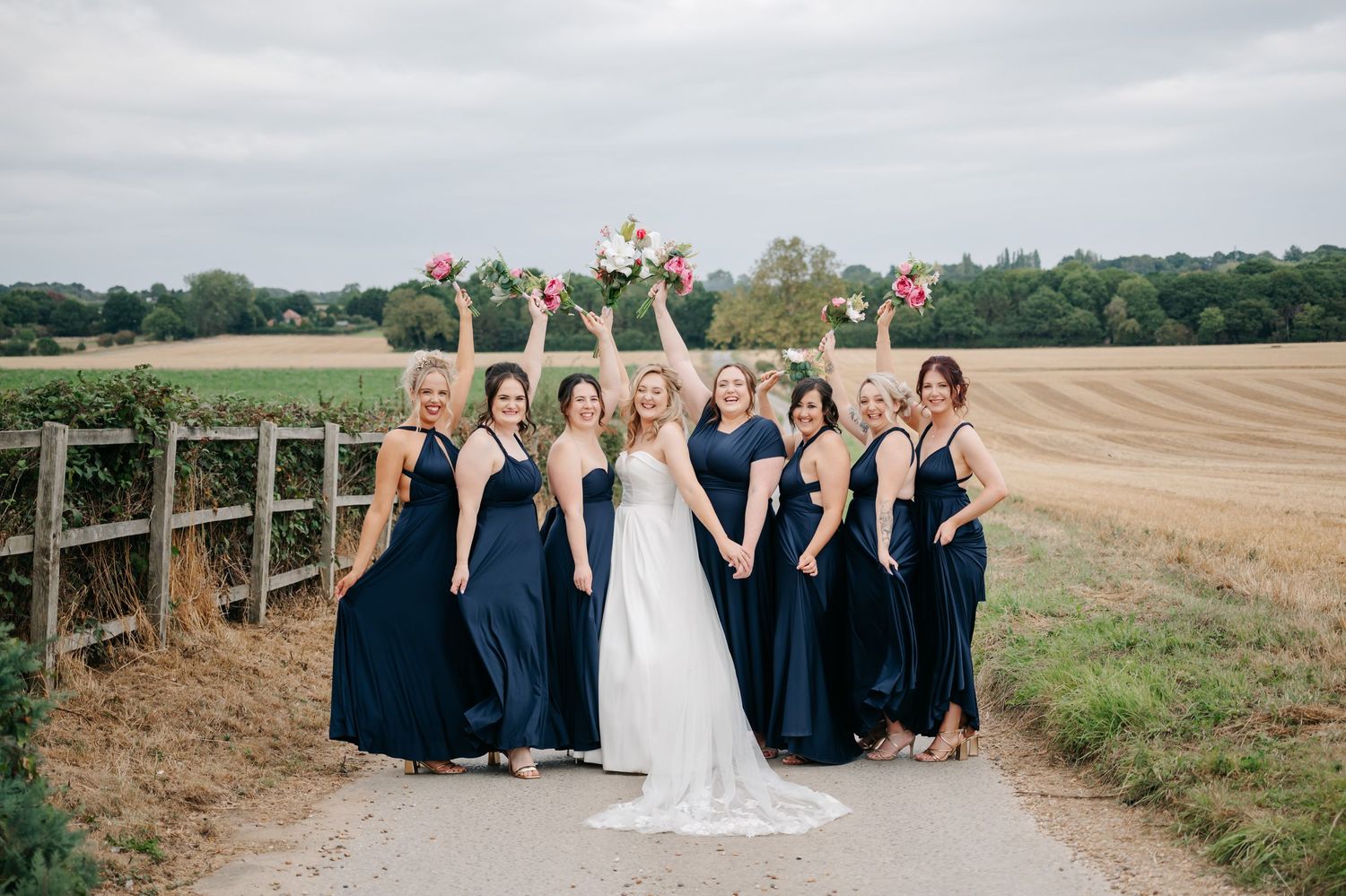 Wedding party in navy blue dresses and suits poses together on rustic wooden bridge in countryside setting.