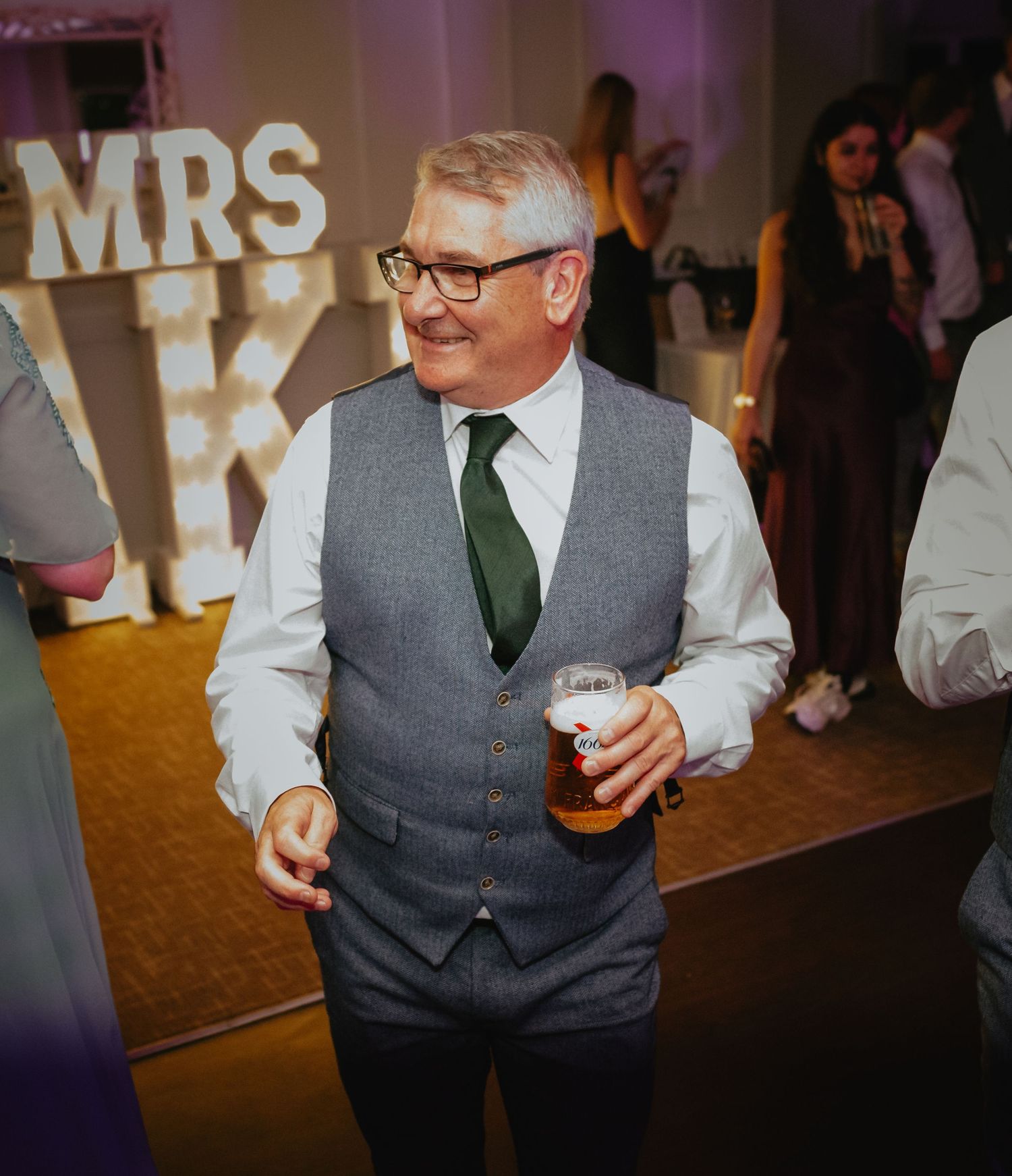 Wedding guests in formal attire mingle at an evening reception with illuminated signage in background.