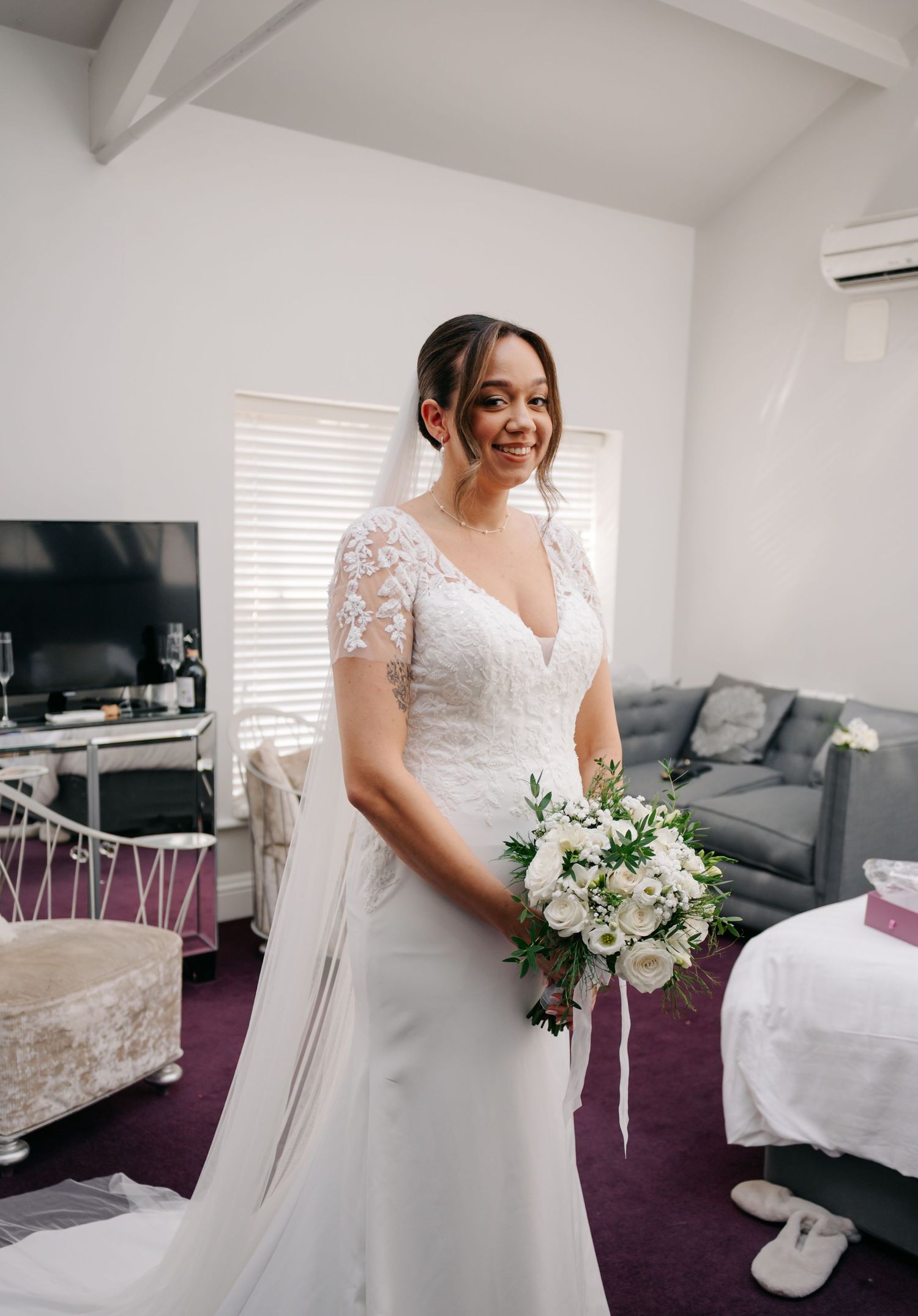 A bride in a white lace wedding dress poses in a modern room with purple carpet and white walls.