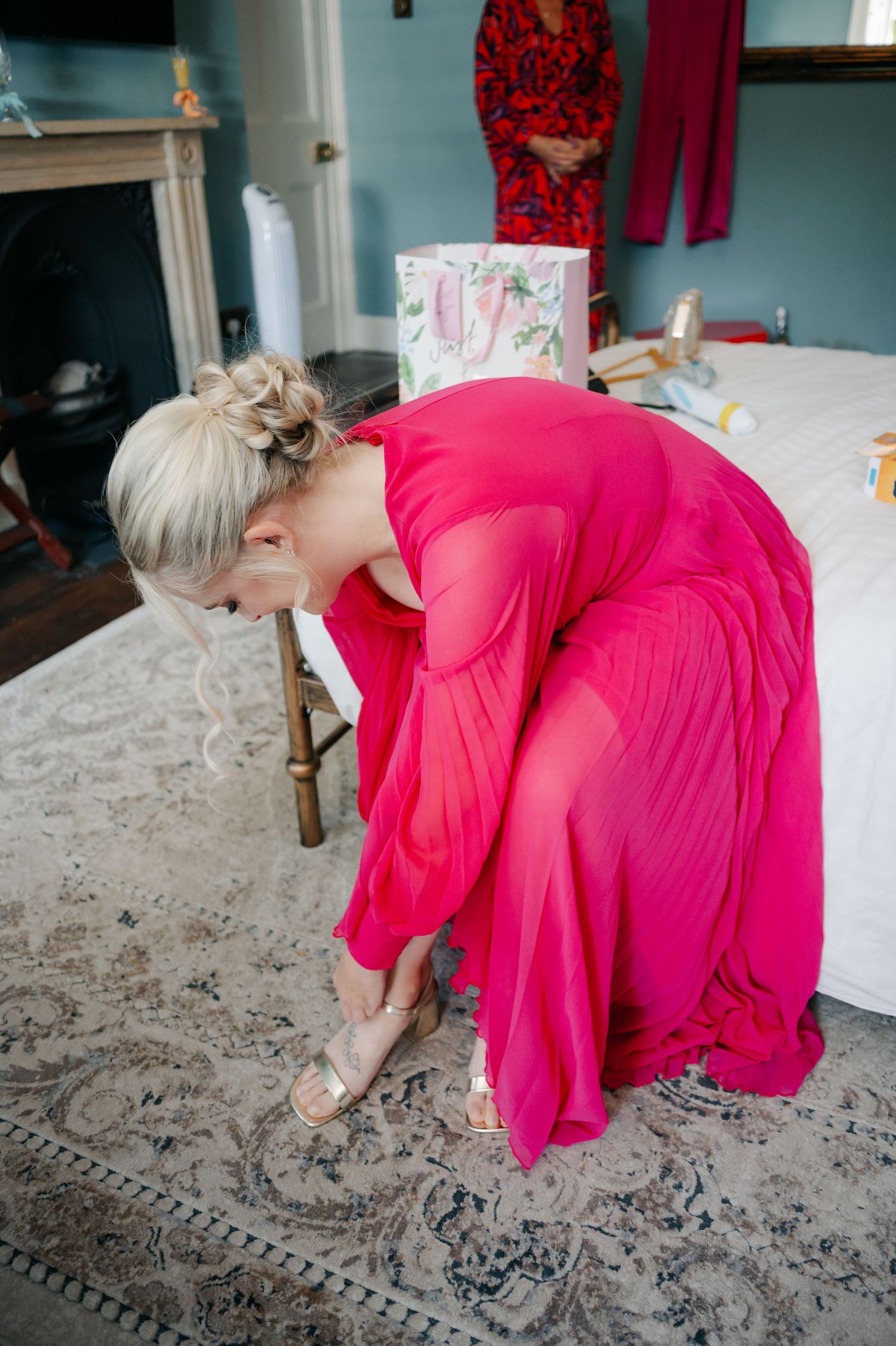 A person in a vibrant pink dress bends down to adjust something on a patterned area rug in a bright room.