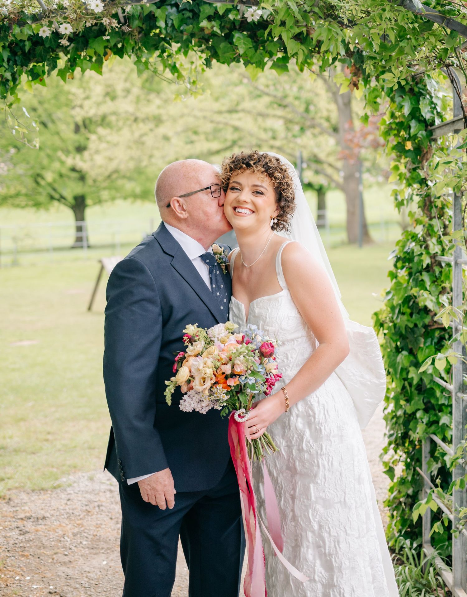 Wedding couple embracing under a lush green archway in an outdoor garden setting.