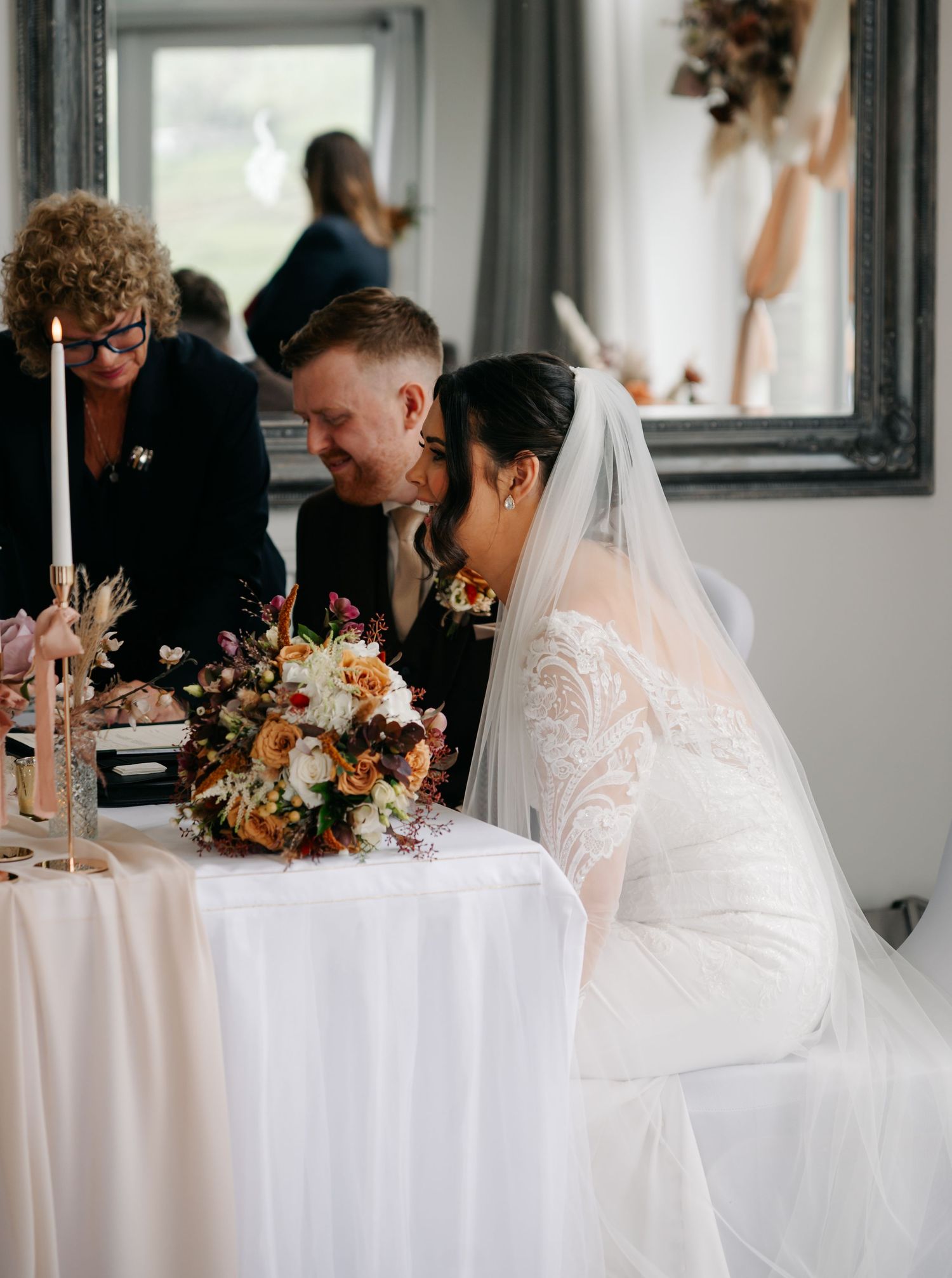 Elegant wedding reception table setup with white linens and autumn-colored floral centerpieces in a bright venue.