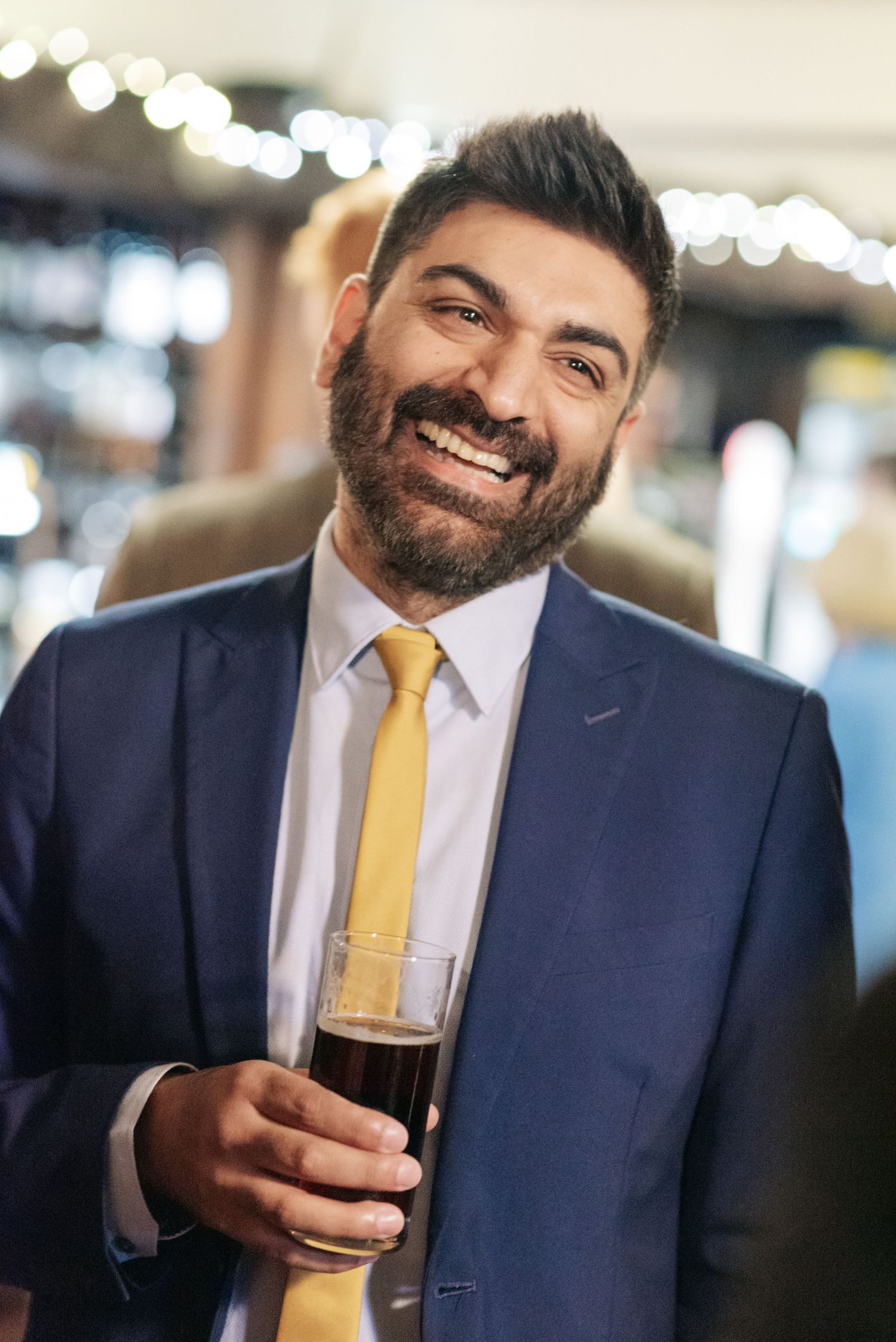 Man in navy suit with yellow tie enjoying a dark beer while smiling at camera.