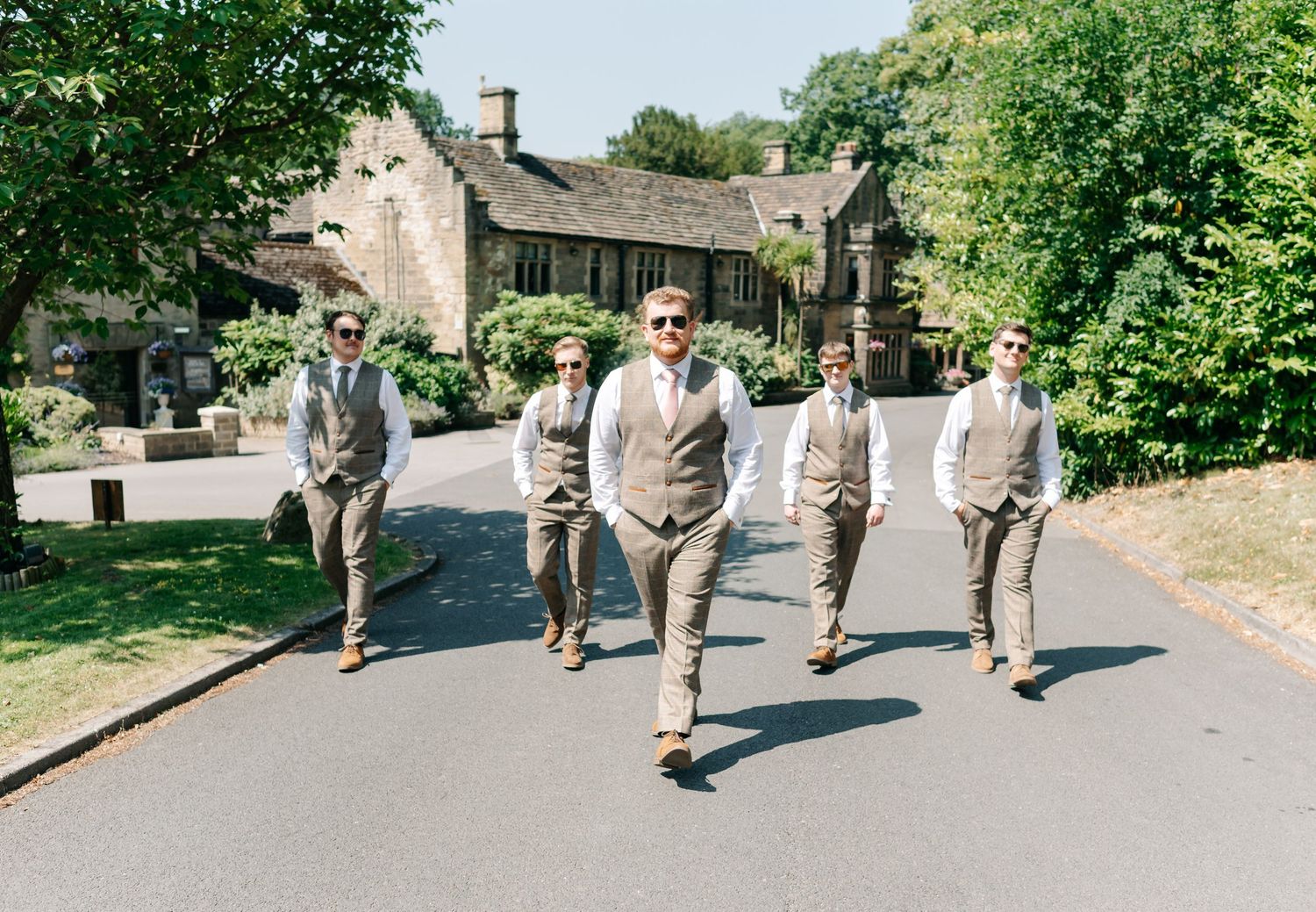 Groom and groomsmen in tan suits walking confidently down a sunlit road at an English countryside wedding venue.