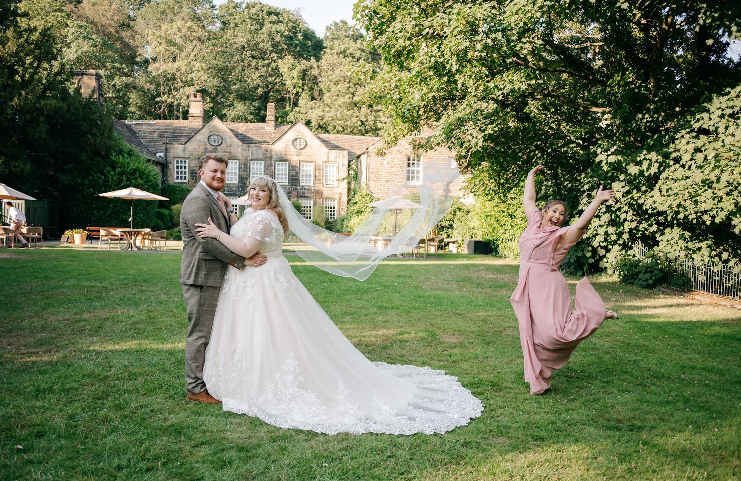 Bride and groom pose in sunny garden as bridesmaid in pink dress twirls beside historic country house venue.