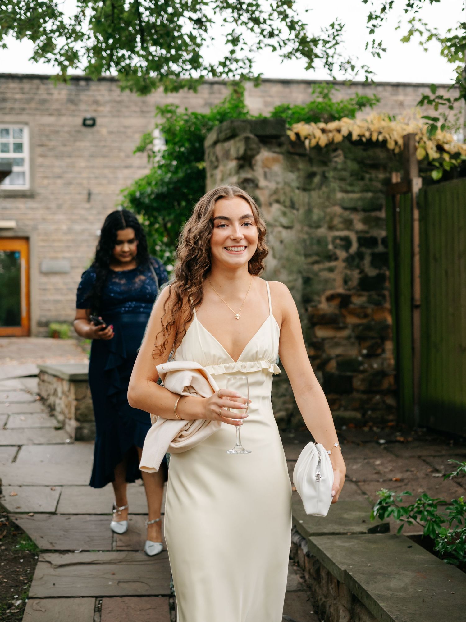 Elegant bride in a fitted silk dress walking outdoors near stone buildings.