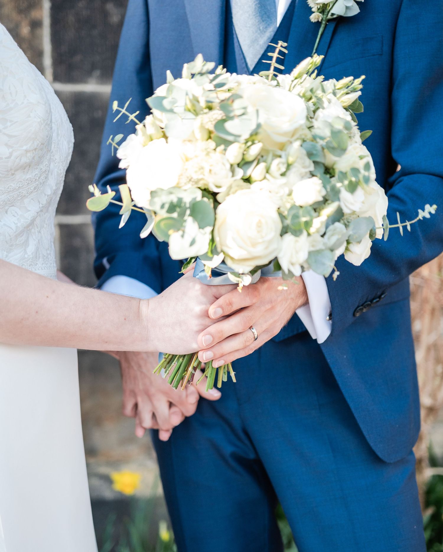 Close-up details of white and green wedding bouquet against navy blue suit during ceremony.