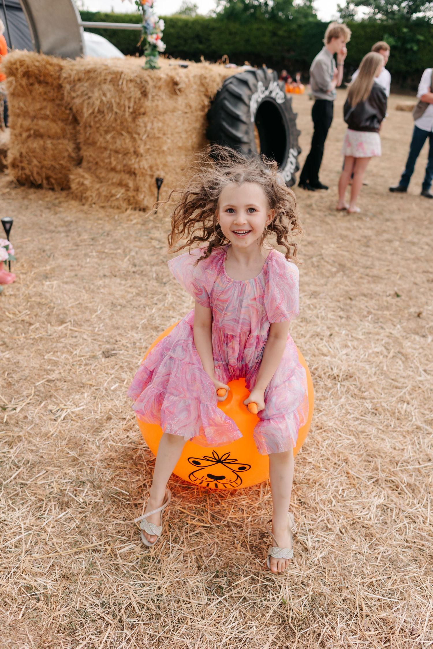 Young guest in pink dress playing on hay-covered ground at outdoor wedding reception.