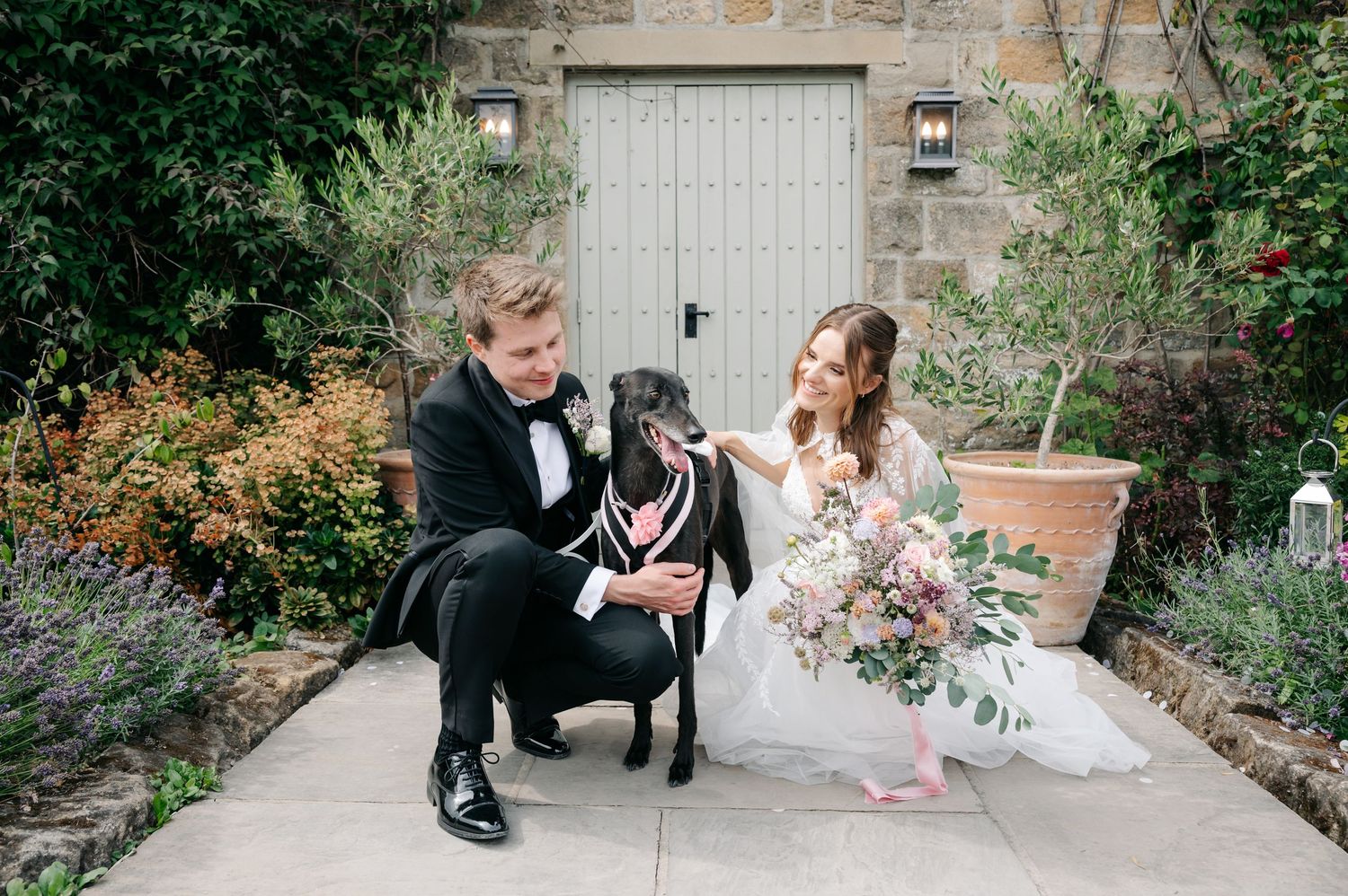 Bride and groom in formal wedding attire kneel beside their black Labrador dog in a lush garden courtyard.