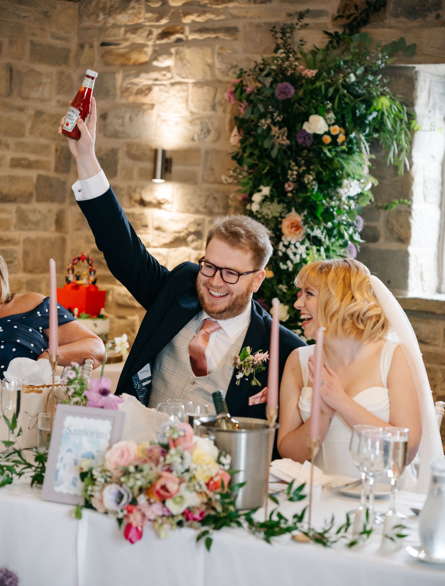 Groom raises a bottle in celebration at wedding reception table with bride laughing beside floral arrangements.