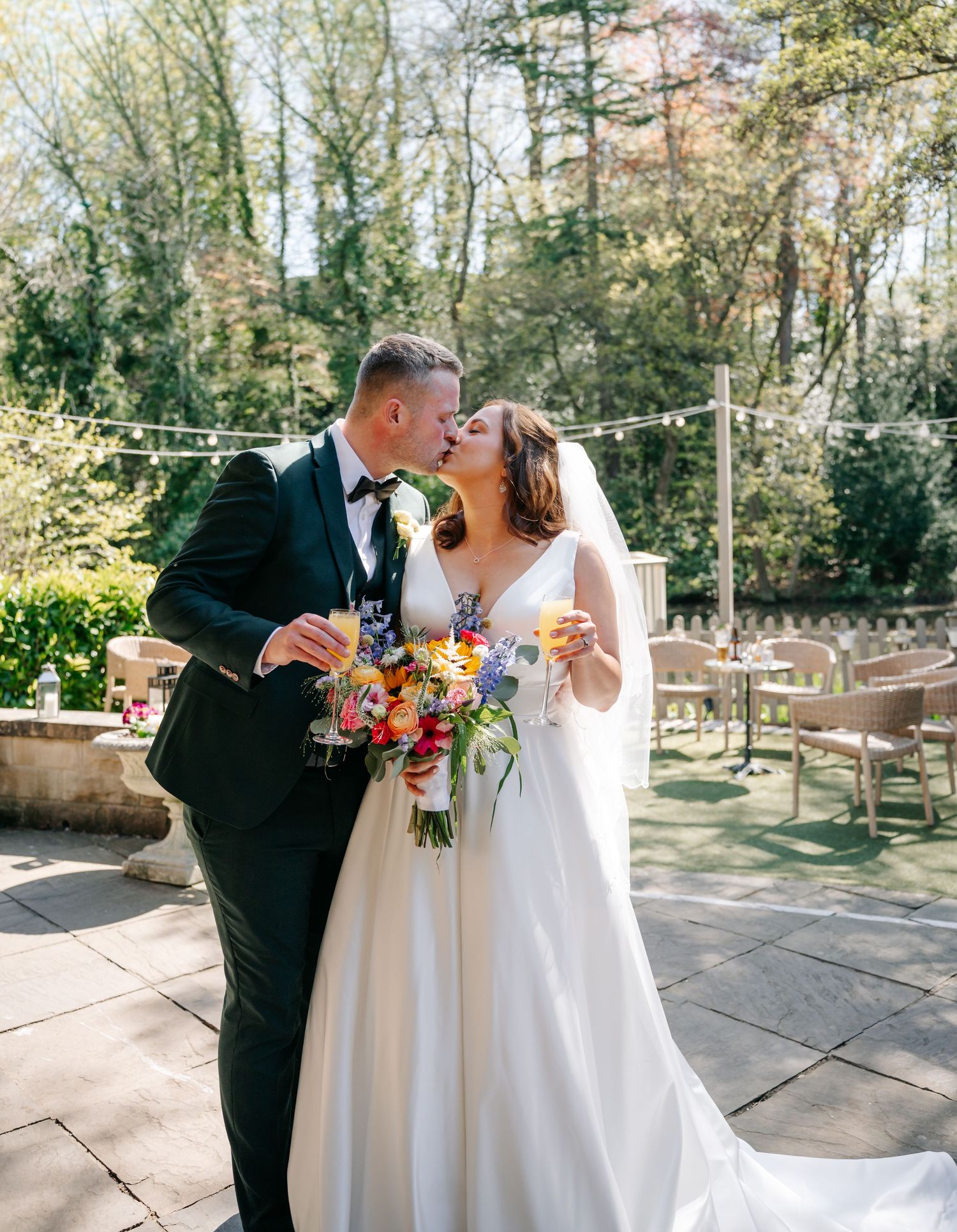 Bride and groom kissing on a sunlit patio, holding champagne glasses, with a colorful bridal bouquet and lush garden backdrop.