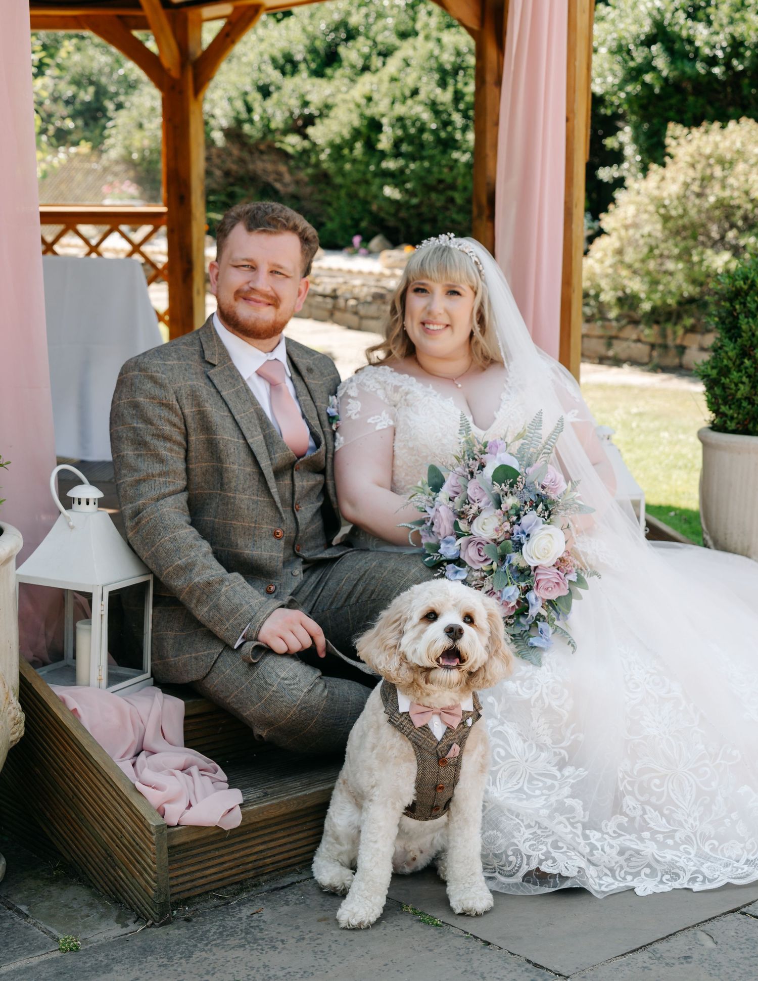 Bride and groom pose with their dressed-up Cavapoo dog under a pink gazebo outdoors.