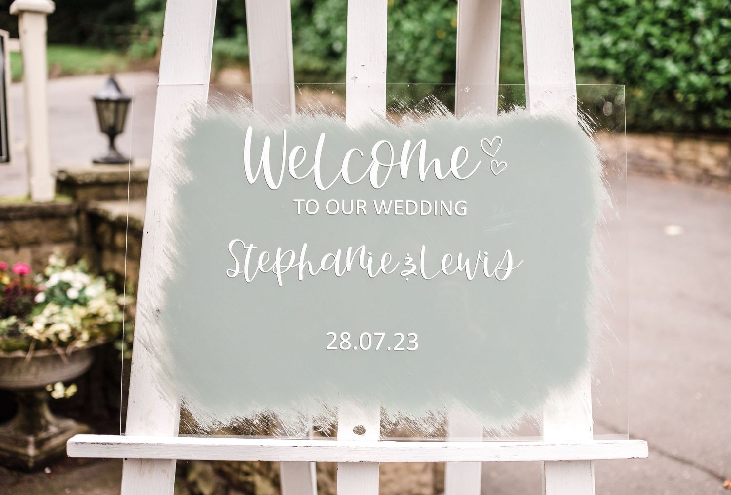A sage green wedding welcome sign displayed on a white stand with the date 28.07.23.