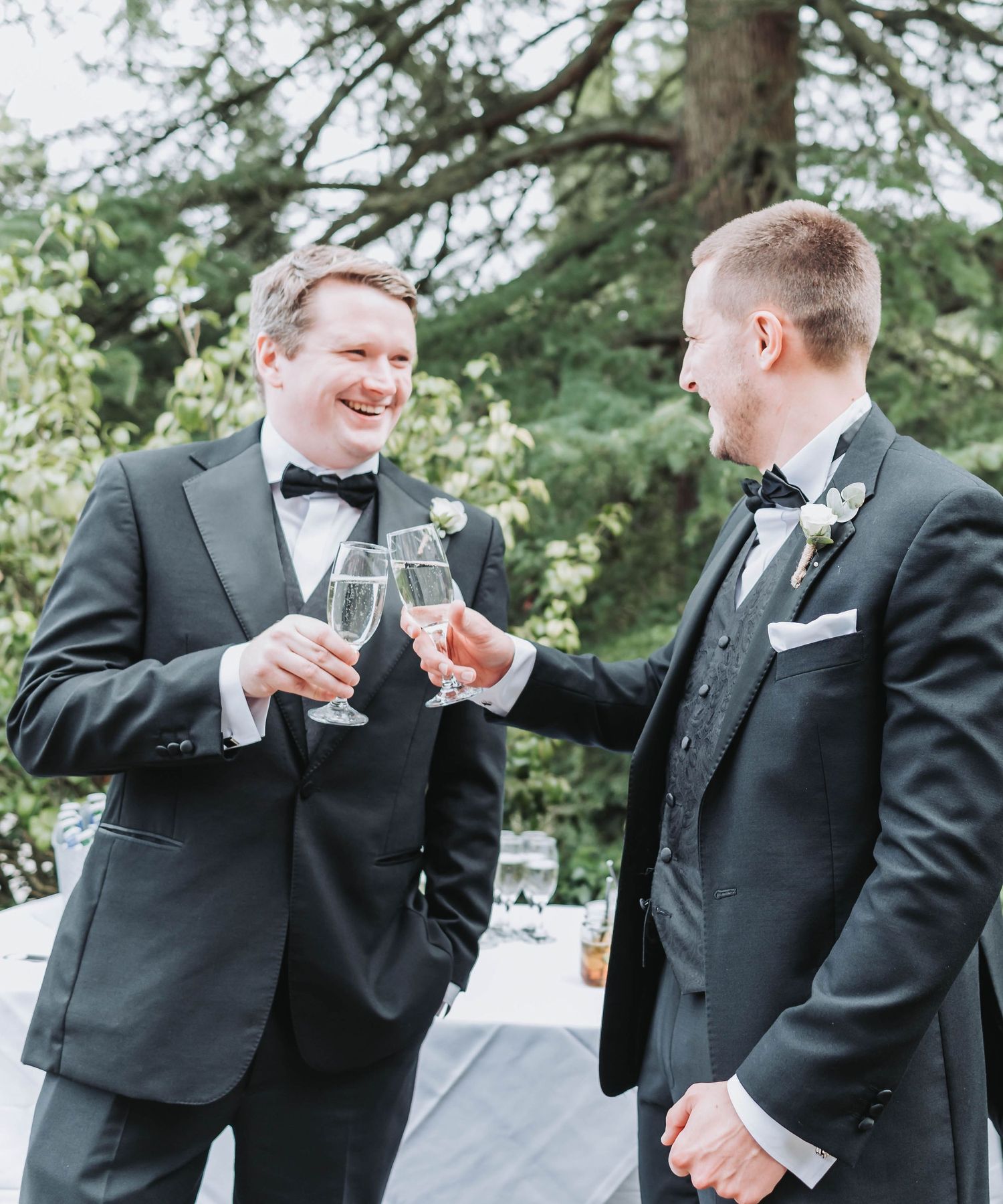 Two men in formal tuxedos toast with champagne glasses outdoors near evergreen trees.