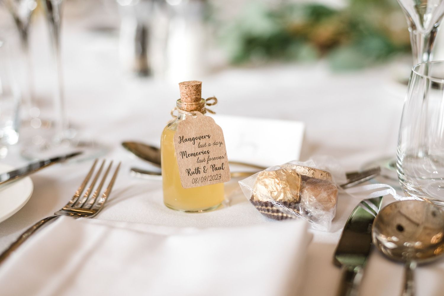 A miniature bottle of honey wedding favor sits beside elegant place settings at a reception table.