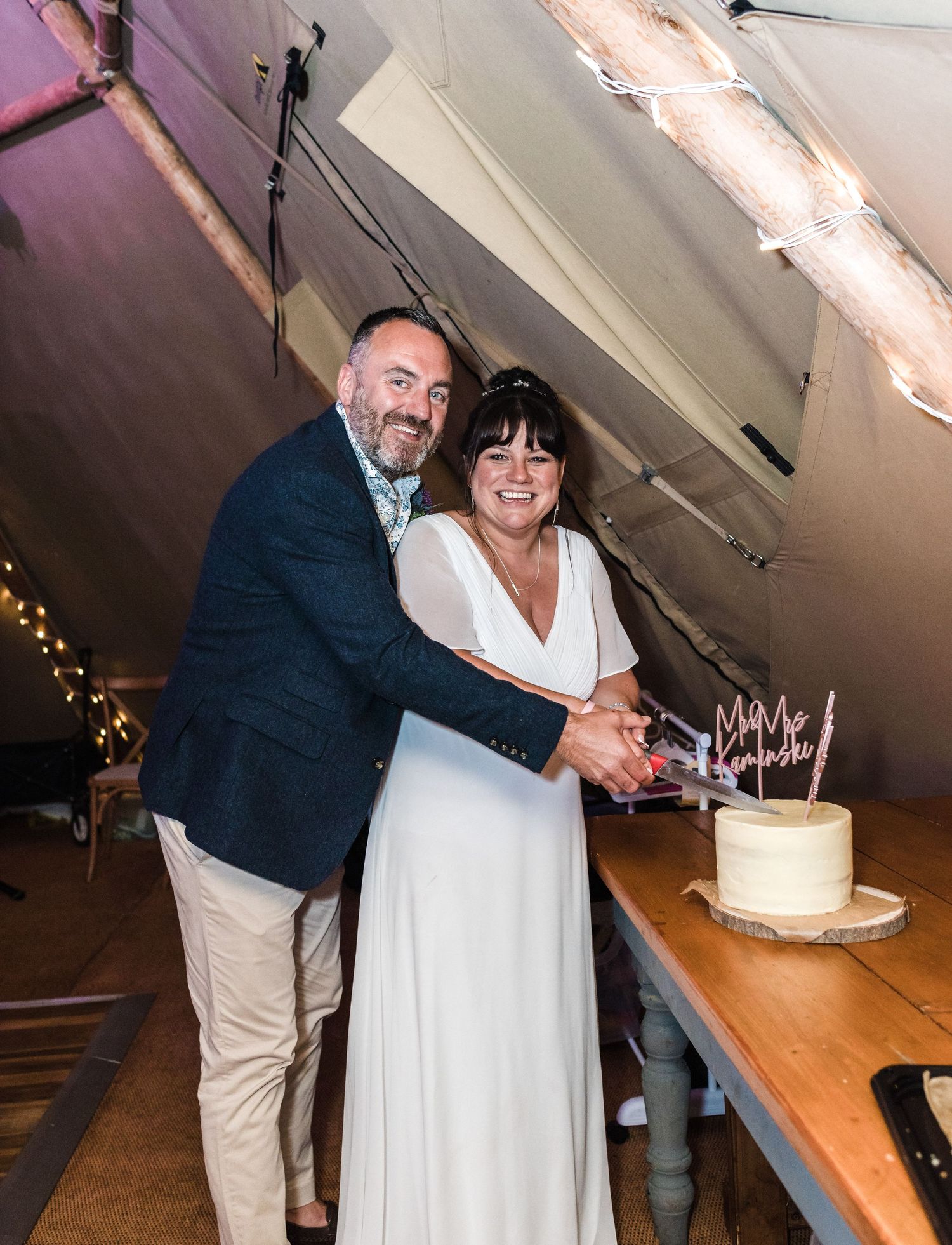 A couple cutting their wedding cake together under string lights in a tent setting.