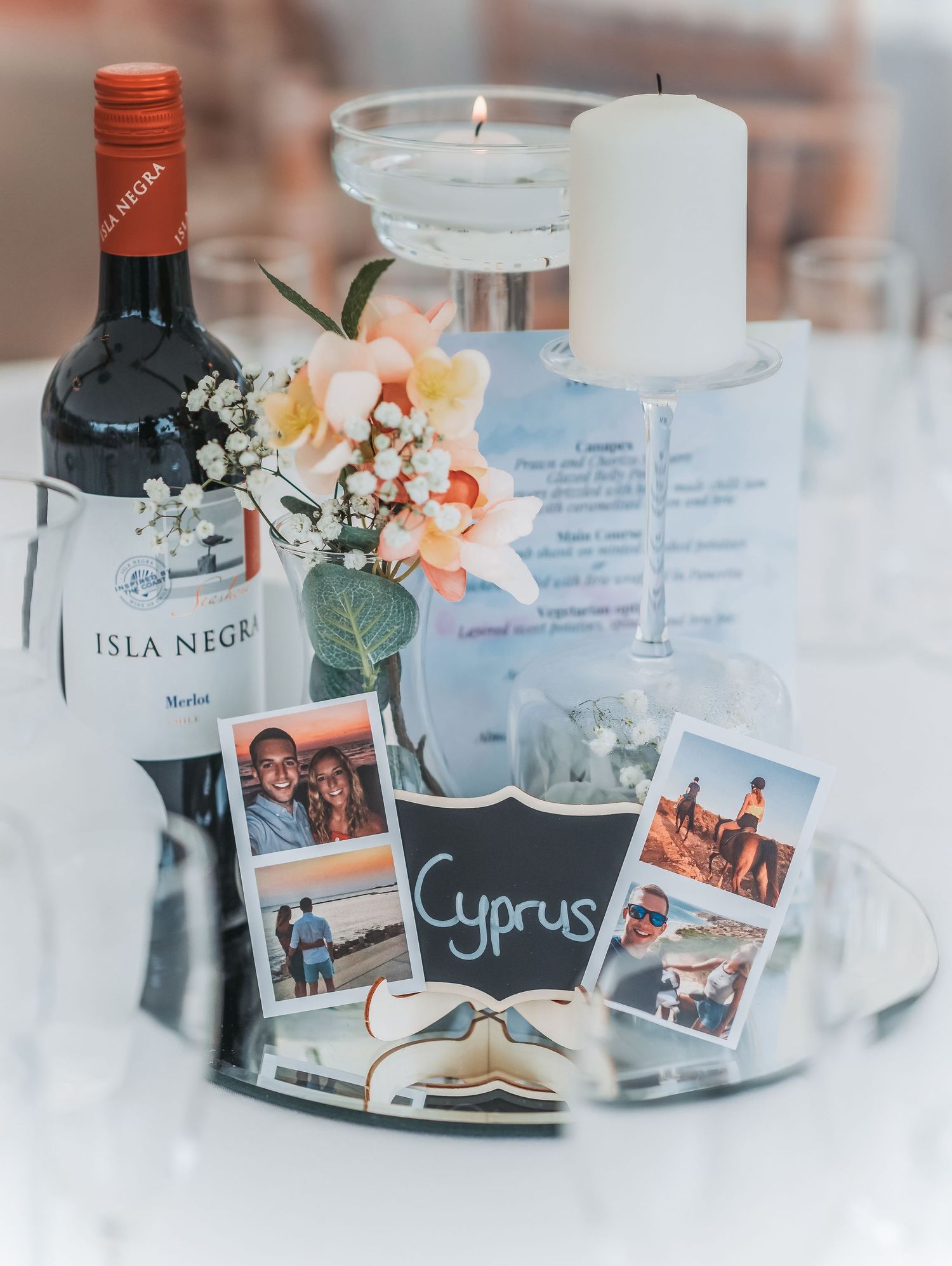 Wedding centerpiece with wine bottle, photos, and Cyprus theme on mirrored tray.