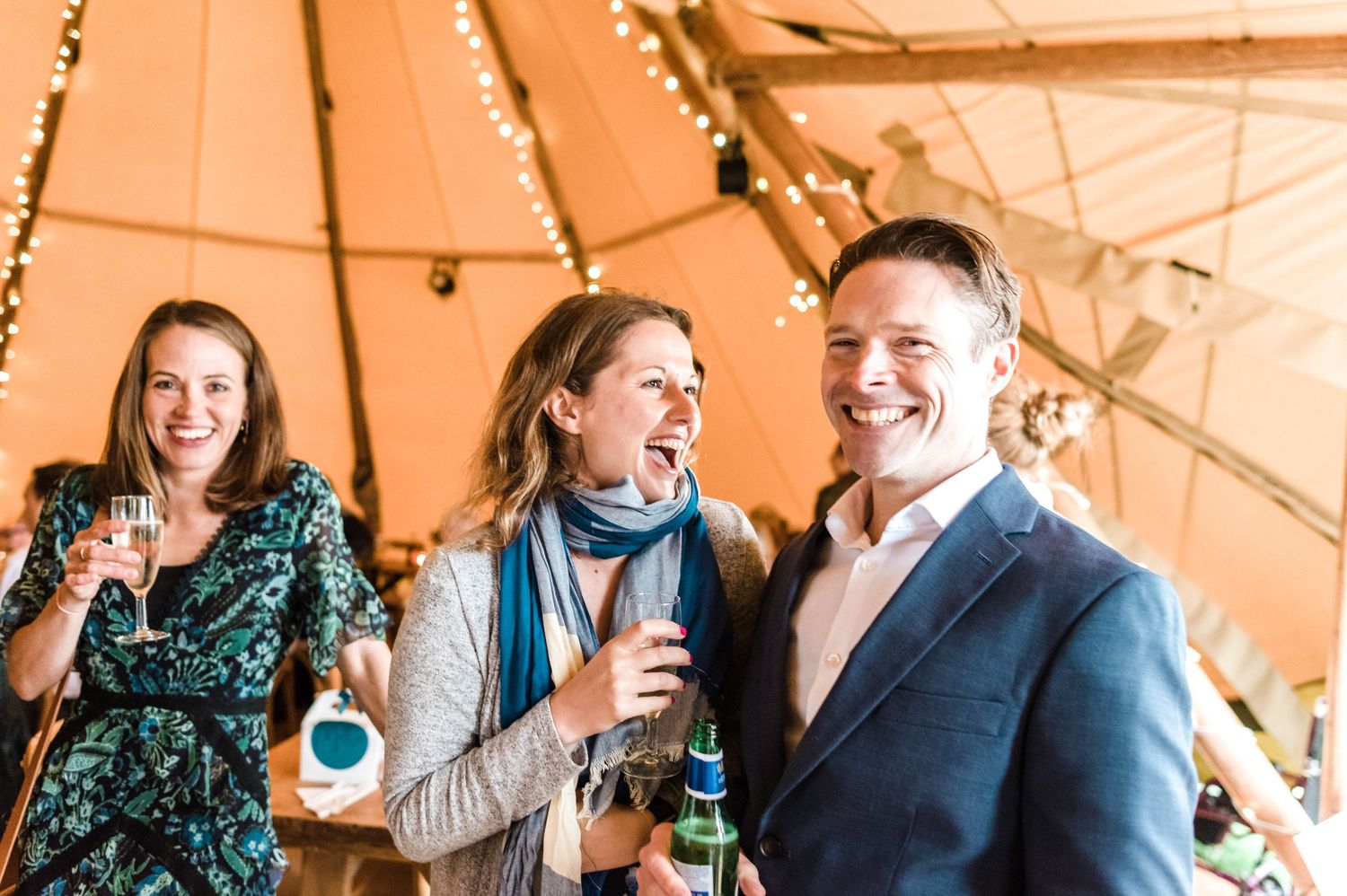Guests enjoying drinks and laughter inside a warm-lit tipi tent reception.