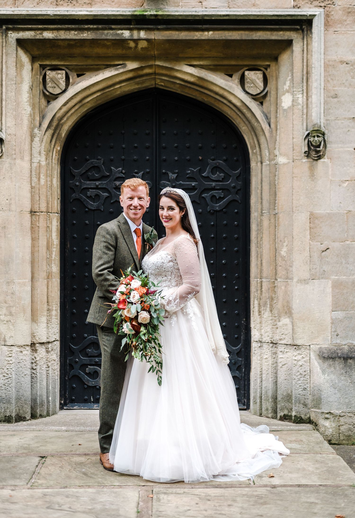 Newlyweds pose in front of an ornate black door at a historic stone church entrance.
