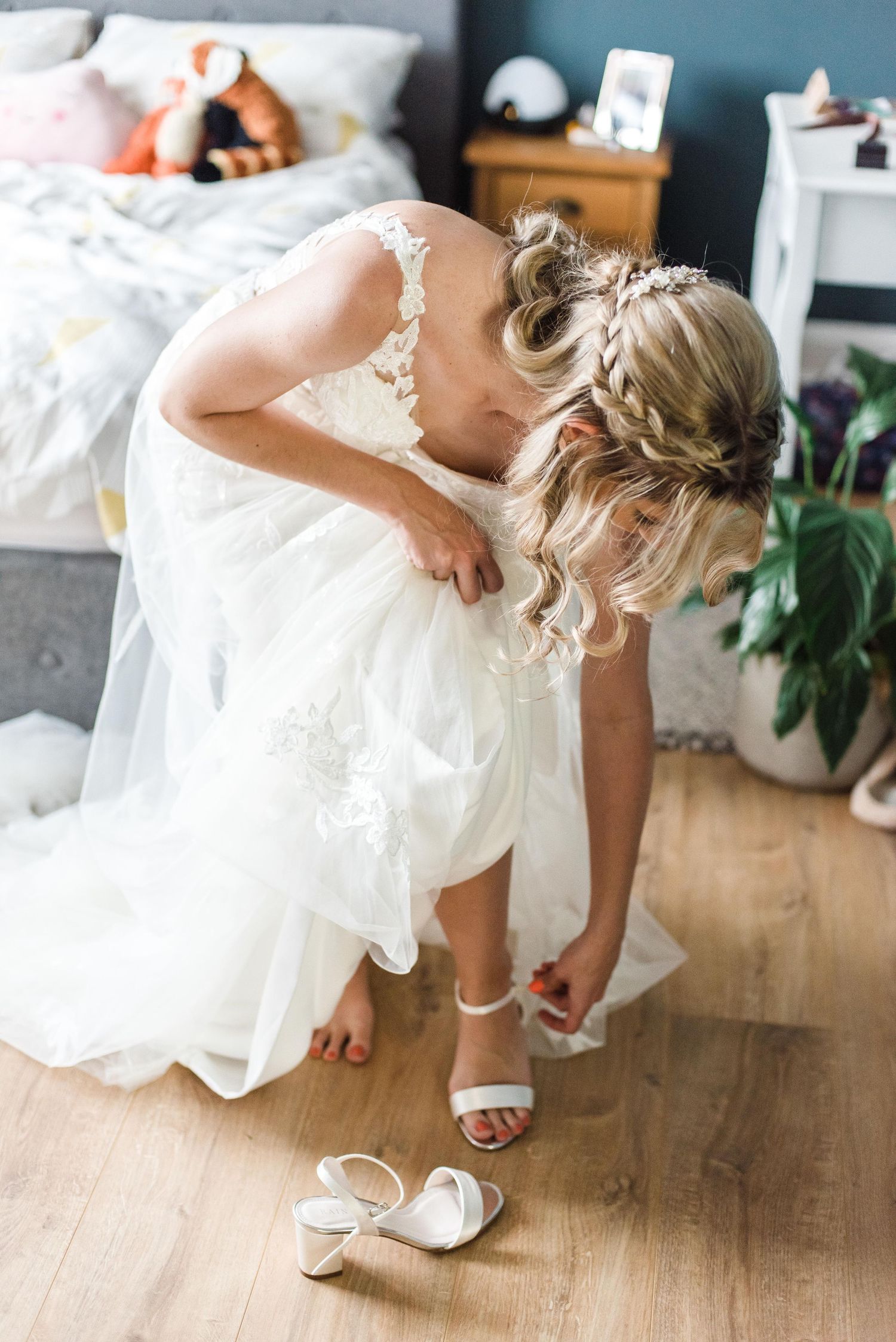 Bride in white lace dress bends down to adjust wedding shoes on wooden floor.