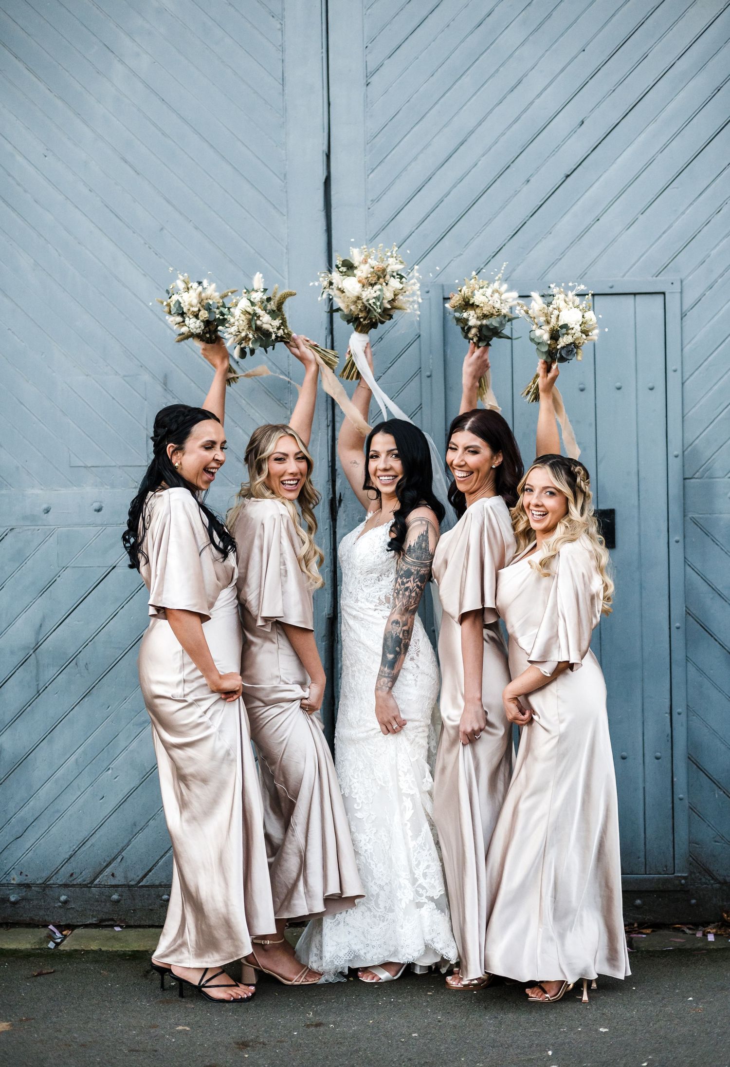 Bridal party in champagne dresses holding bouquets against blue wooden wall.