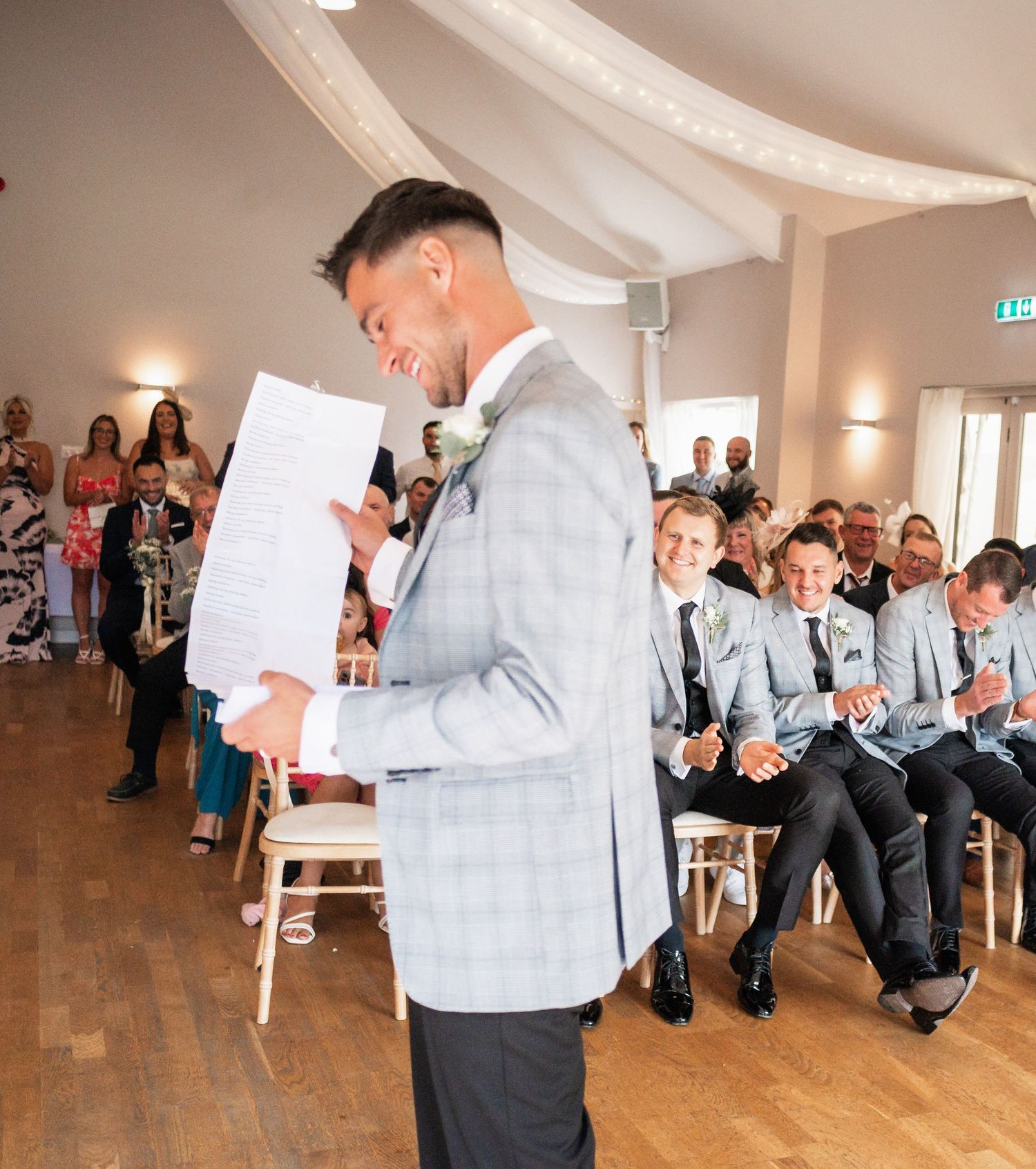 A groom in a light gray suit reads from a paper during an indoor wedding ceremony.