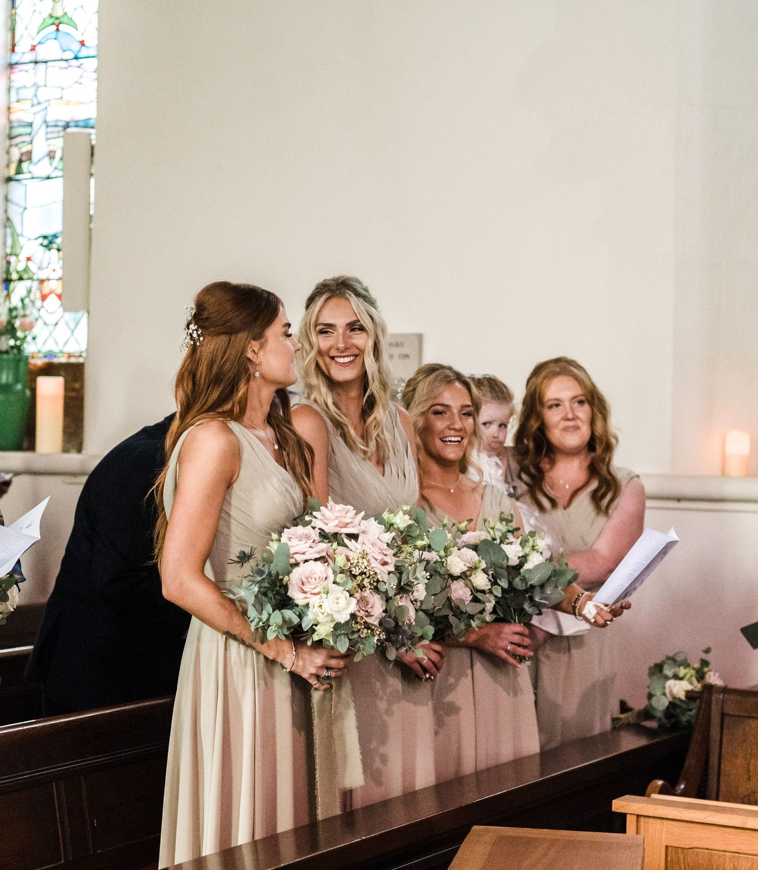 Bridesmaids share a candid moment of laughter while holding pink and white bouquets in a church pew.