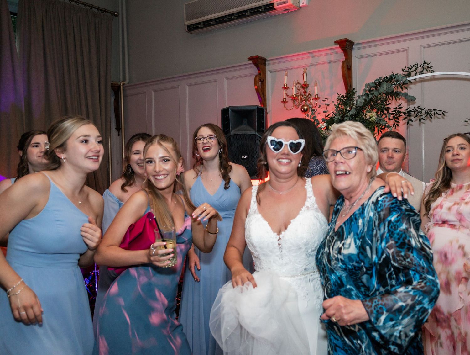 Wedding party dancing together during reception with colorful lighting and festive atmosphere.