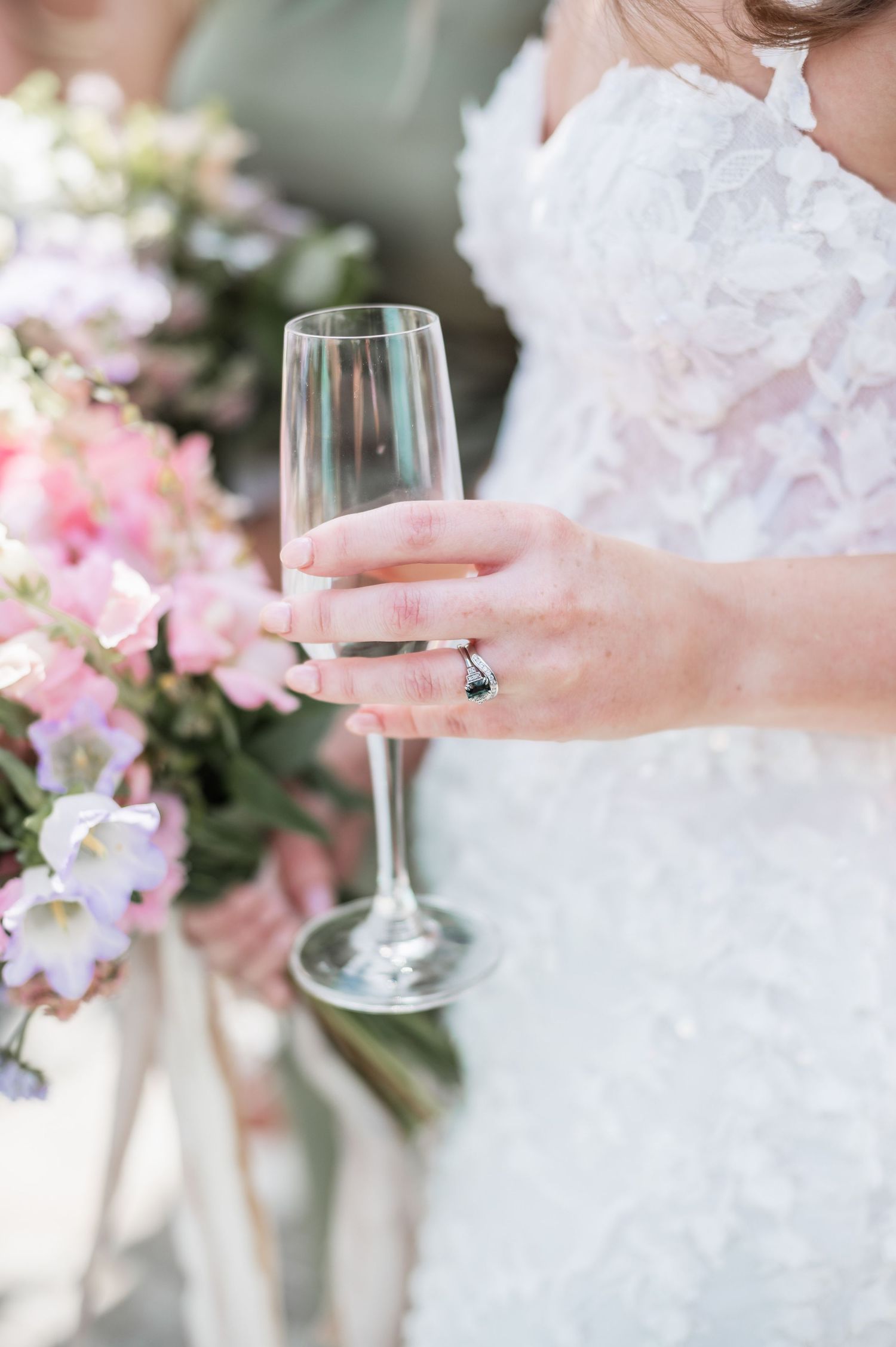Close-up of a champagne glass held by hands with an engagement ring against pink floral decorations.