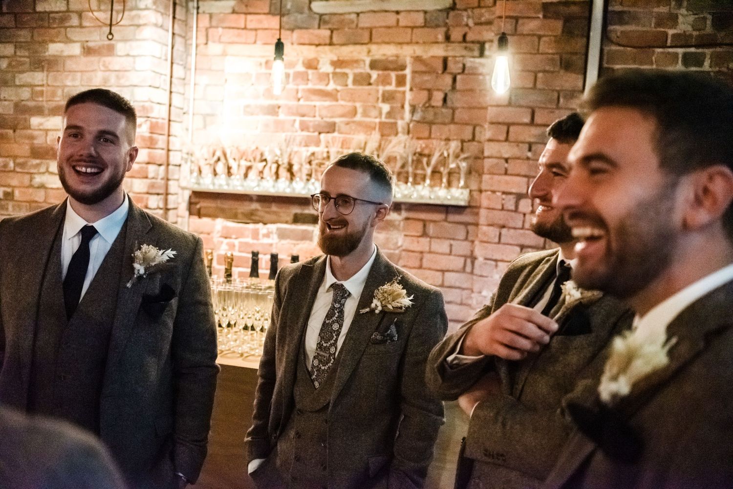 Three men in dark suits and boutonnieres laugh together at wedding reception.