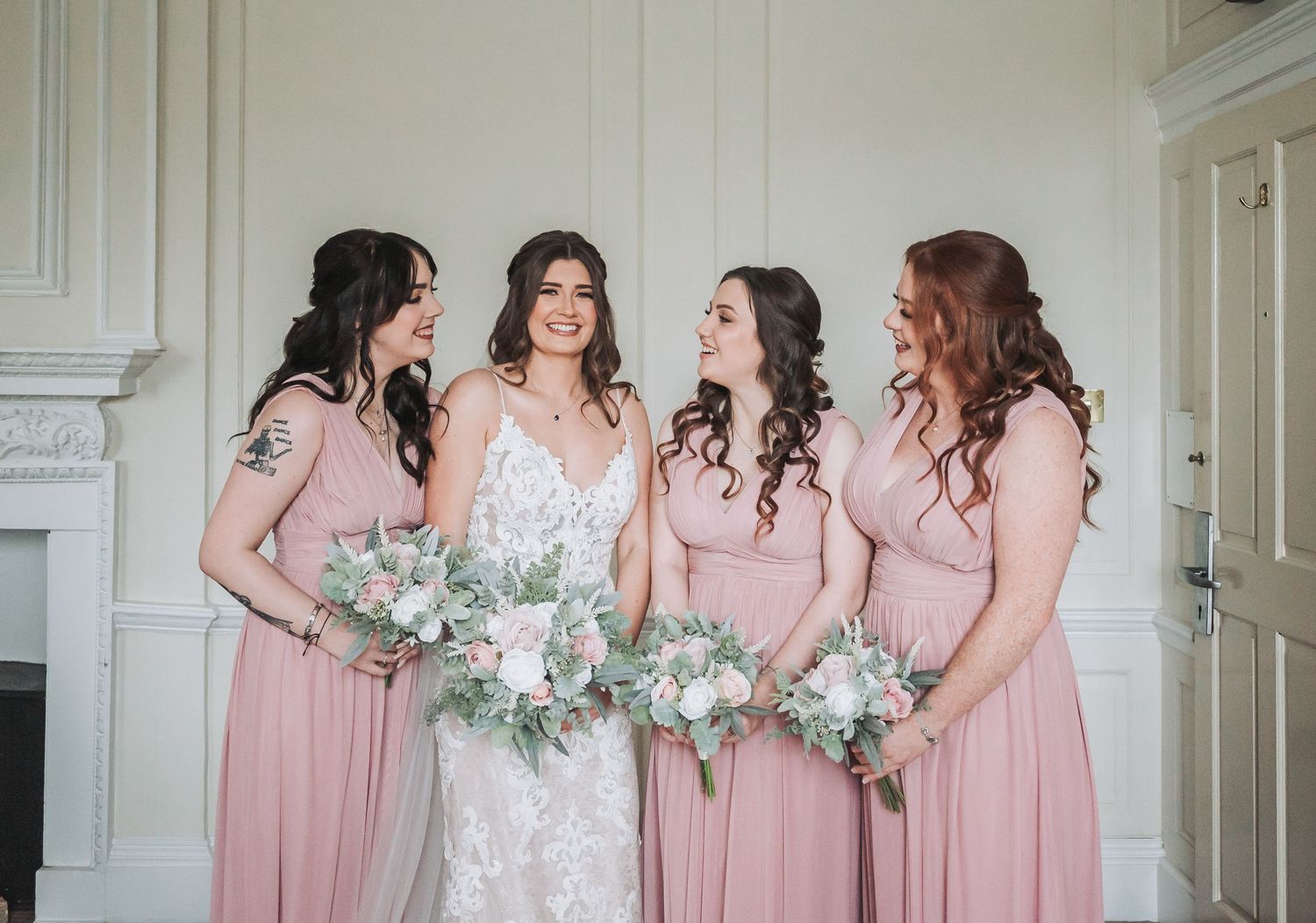 Bridal party in blush pink dresses holding white and green bouquets in an elegant white room.