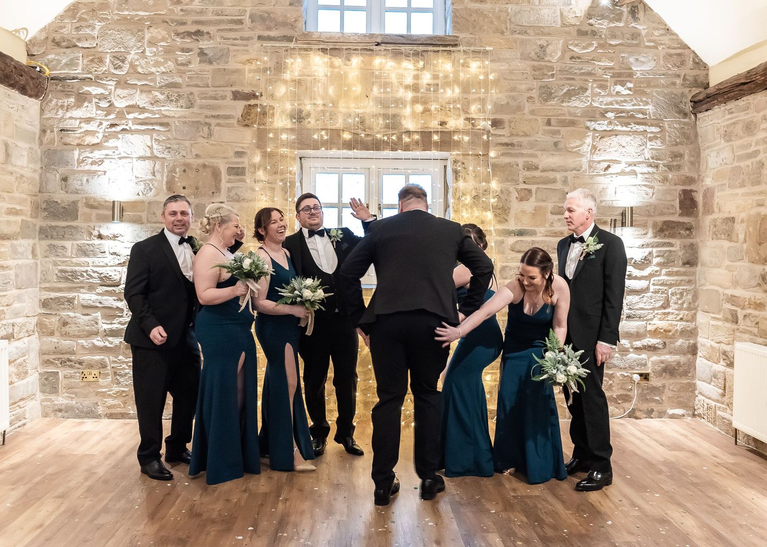 Wedding party in teal dresses and black tuxedos posing in rustic stone venue.