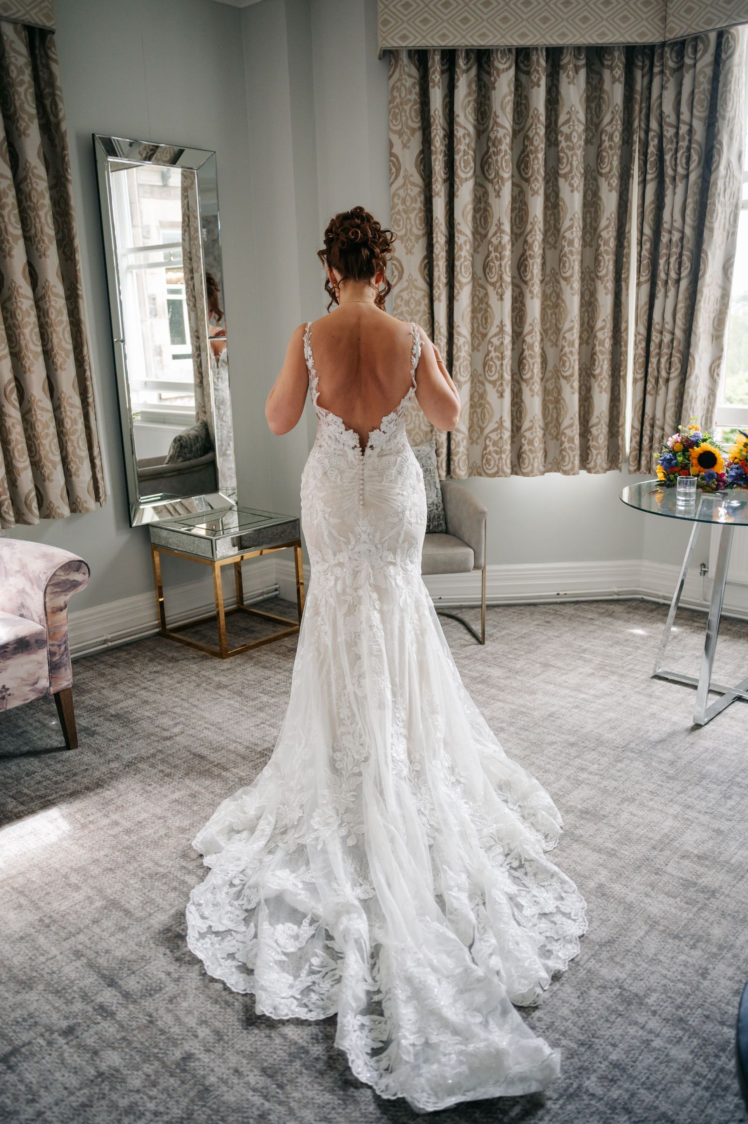 A white lace wedding dress with a dramatic train photographed from behind in a hotel room.